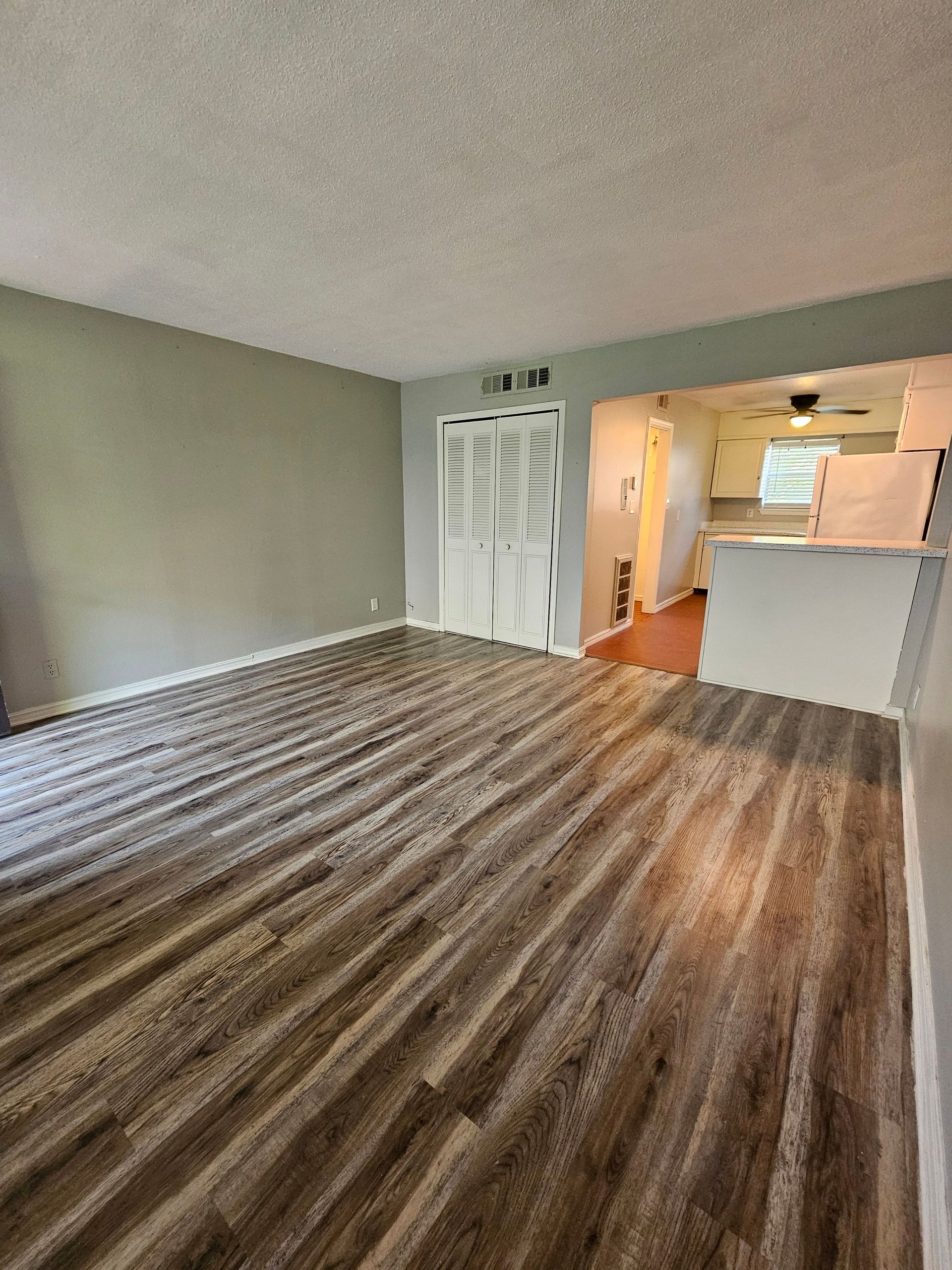 A living room with hardwood floors and a kitchen in the background.