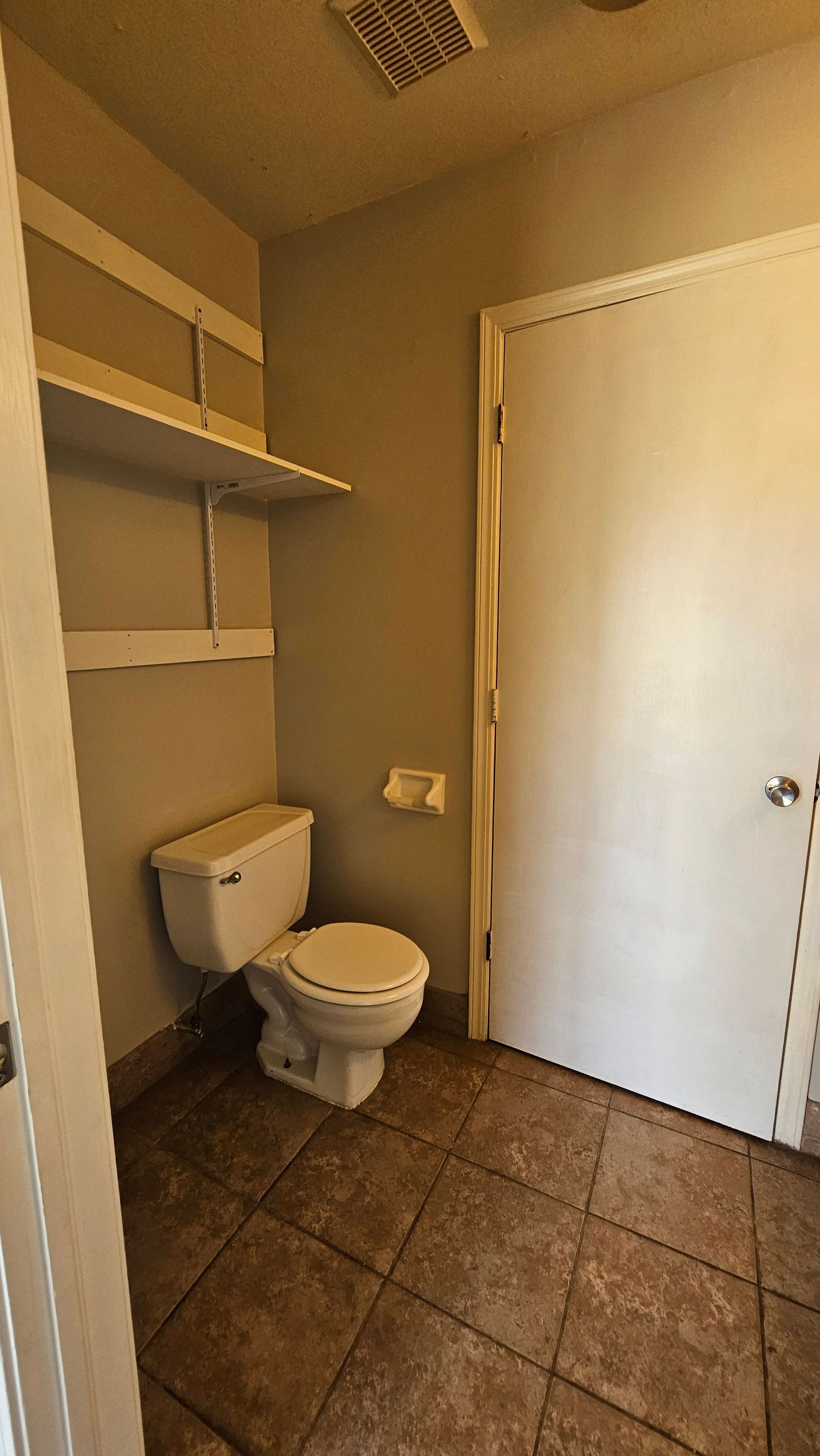 A bathroom with a toilet , shelf , and door.