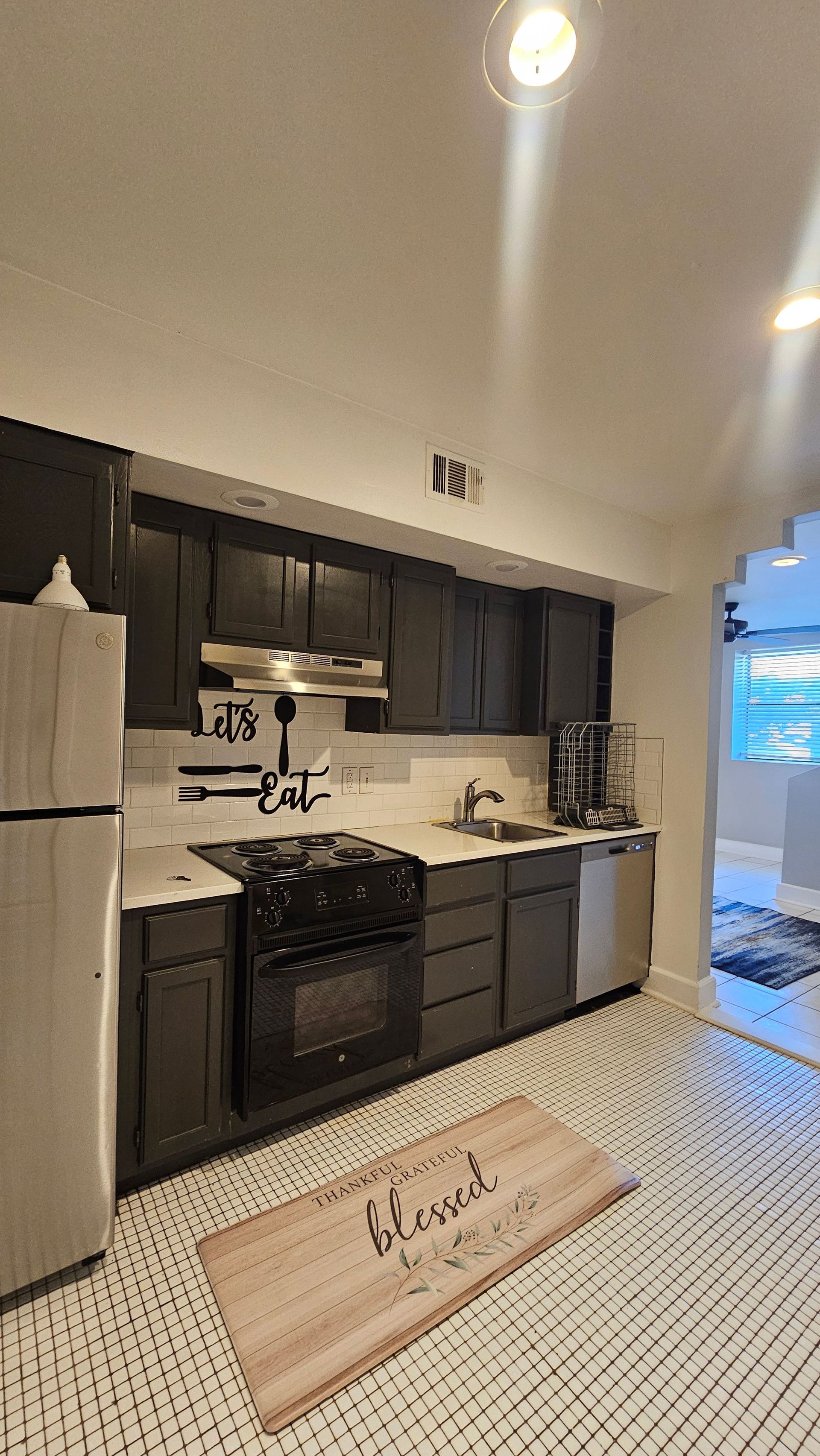 A kitchen with black cabinets , stainless steel appliances , a refrigerator , and a rug on the floor.