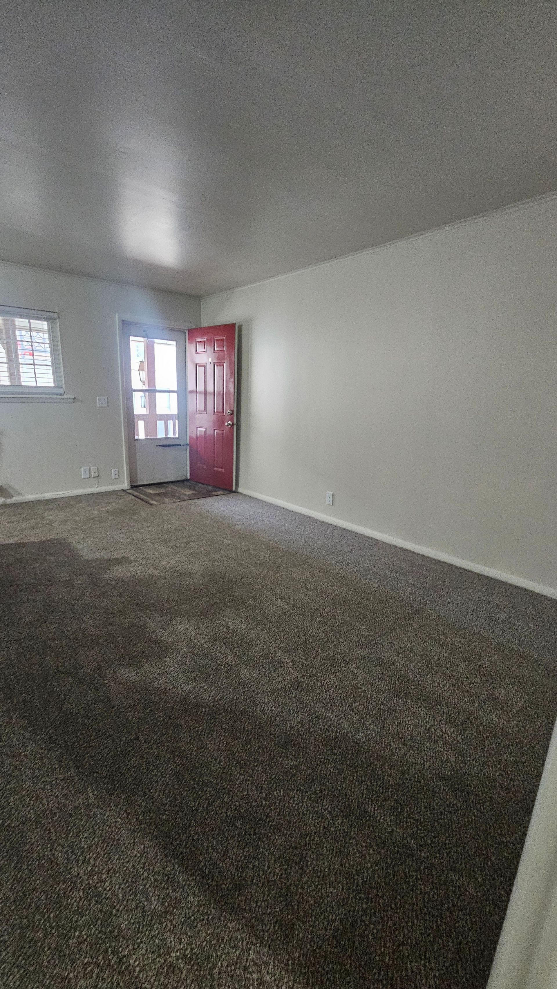 An empty living room with a red door and a window.