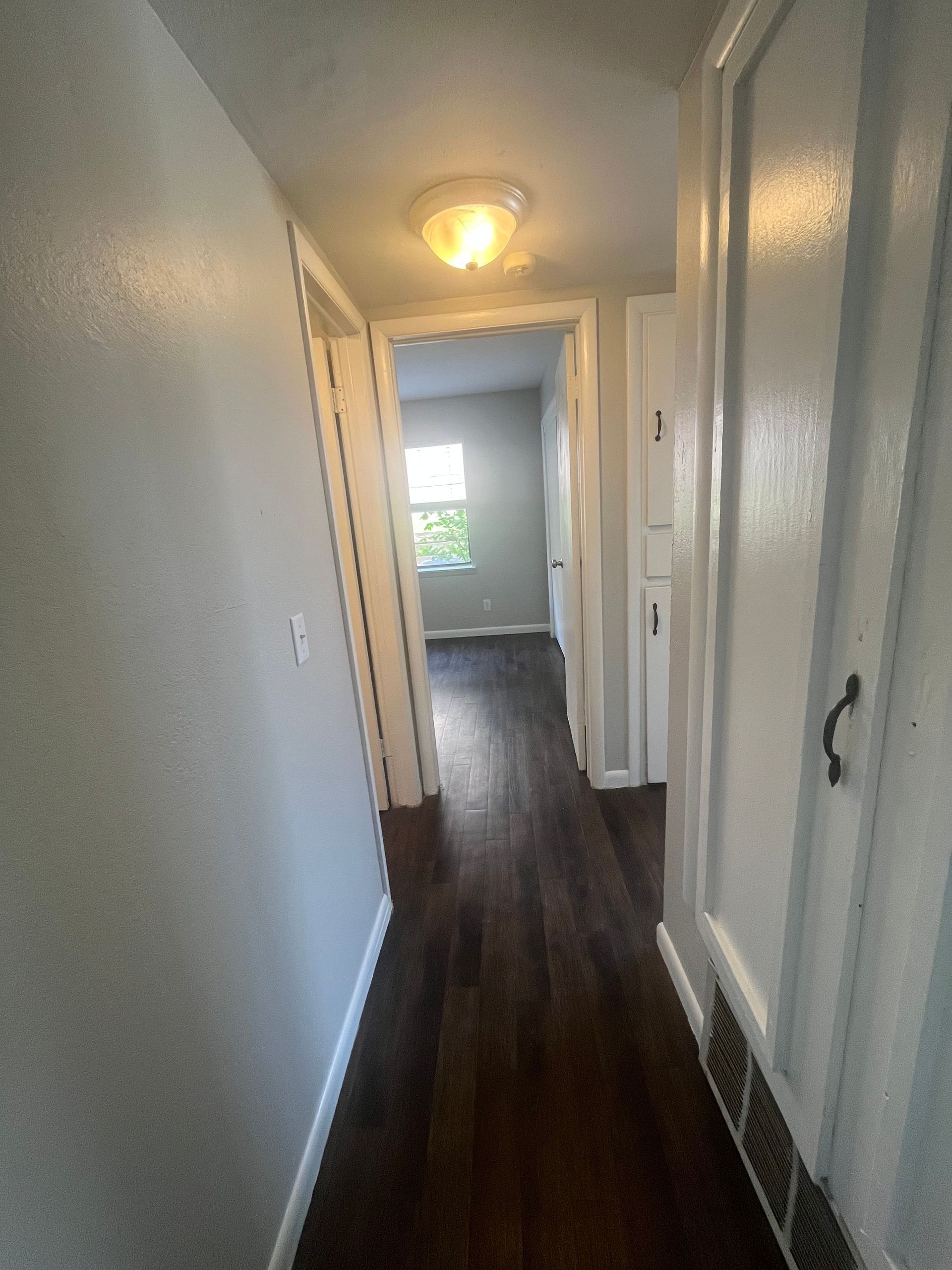 A hallway with hardwood floors and white walls leading to a bedroom.
