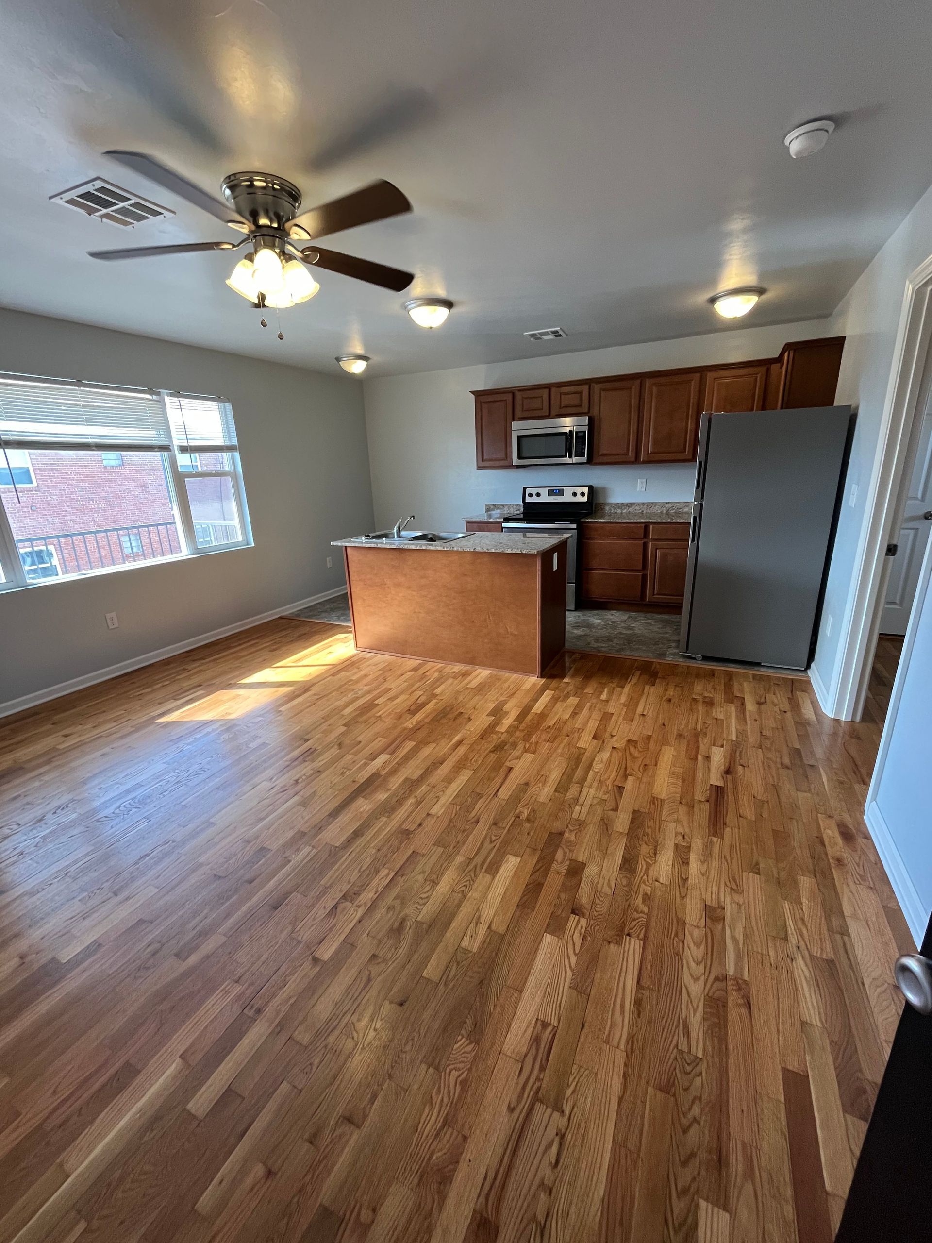 A living room with hardwood floors and a kitchen with a ceiling fan.