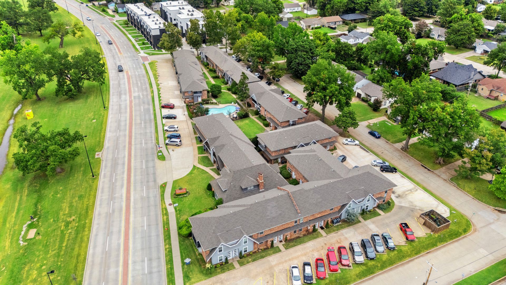 Aerial view of apartment complex with cars parked outside, adjacent to a road with greenery.