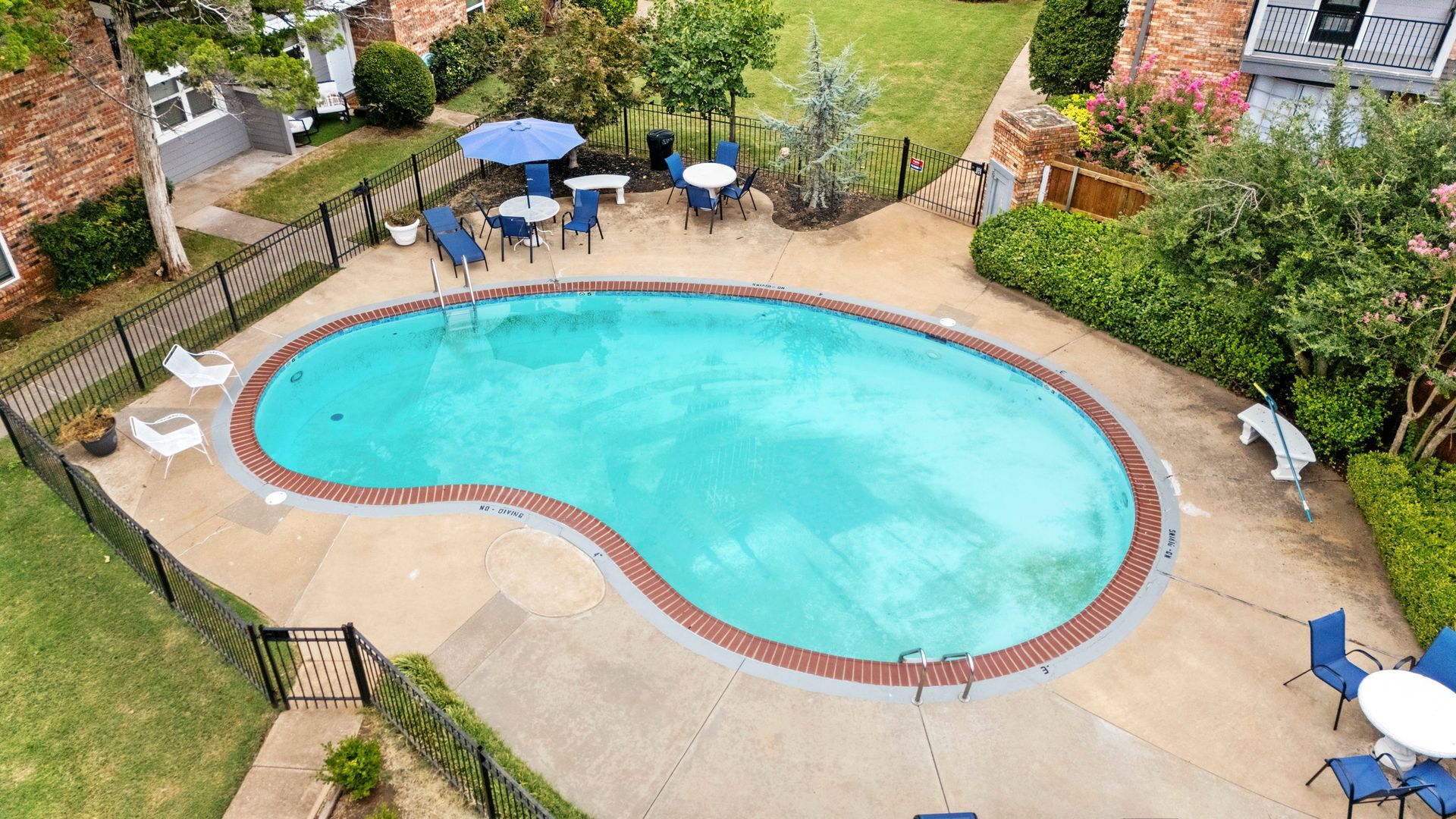 Overhead view of a kidney-shaped pool with surrounding tables, chairs, and shrubbery.