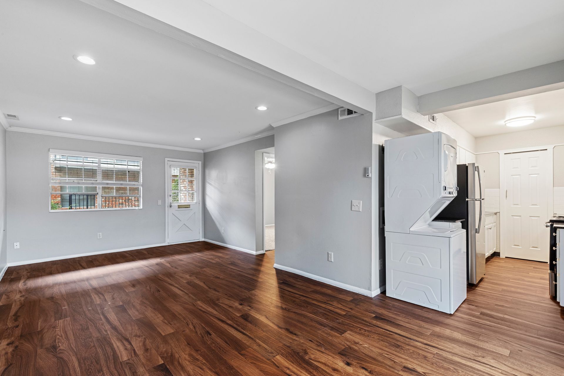 Empty living room and kitchen. Dark wood floors, light gray walls, white cabinets, and stainless steel appliances.
