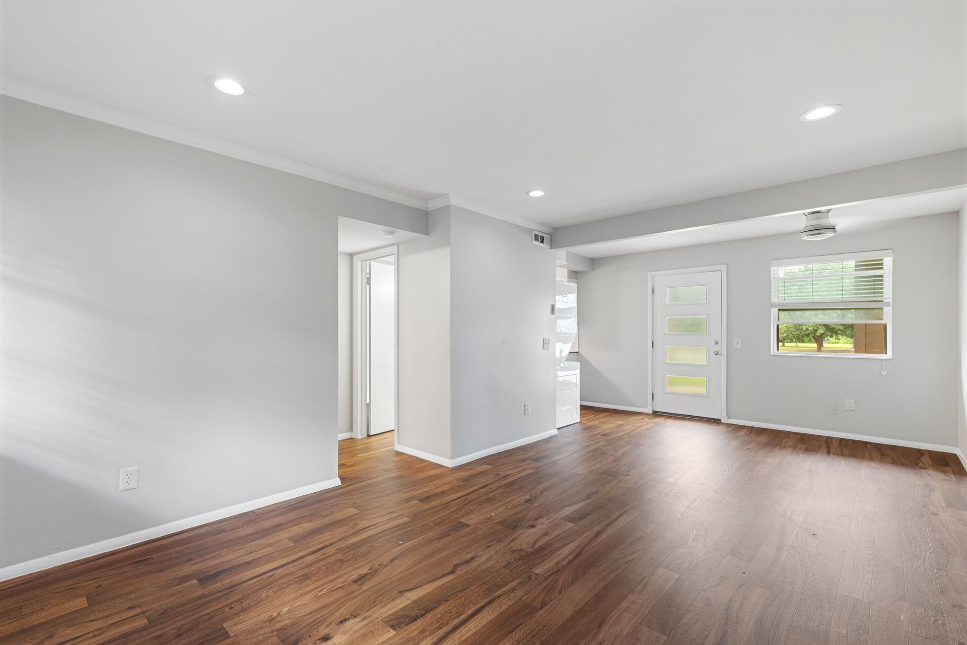 Empty living room with wood flooring, light gray walls, and white trim. Door and window provide natural light.