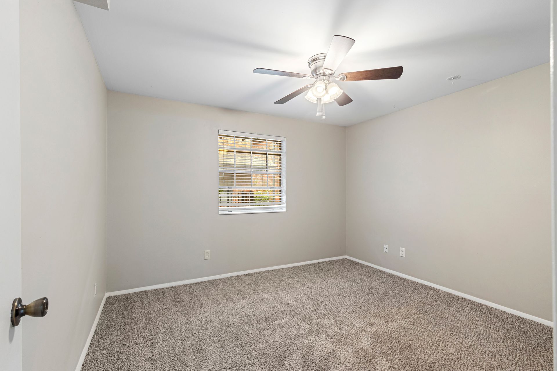 Empty bedroom with neutral walls, carpet, and ceiling fan; a window is visible.