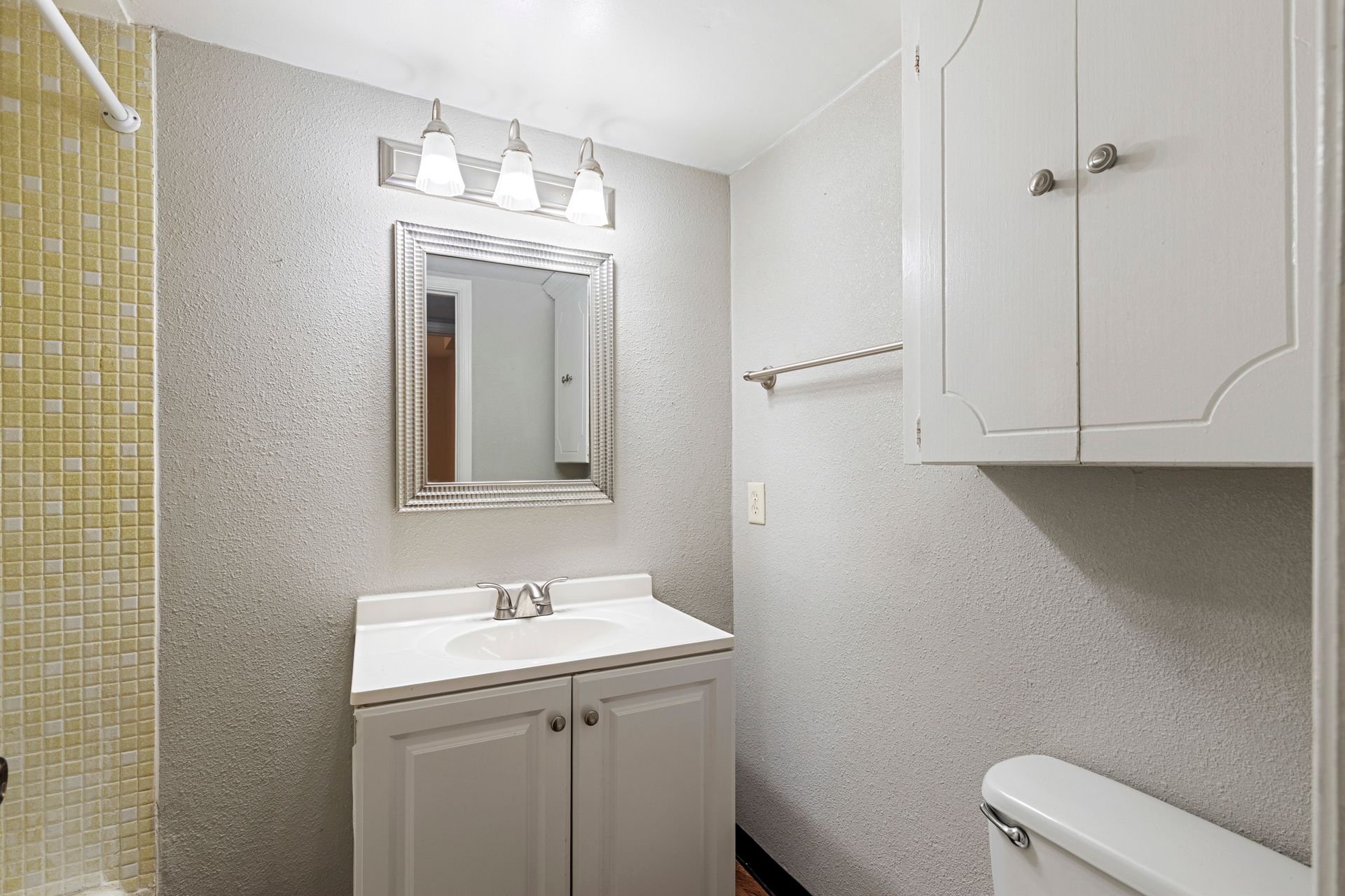 Bathroom with white vanity, medicine cabinet, and toilet; gray textured walls; yellow tiled shower.