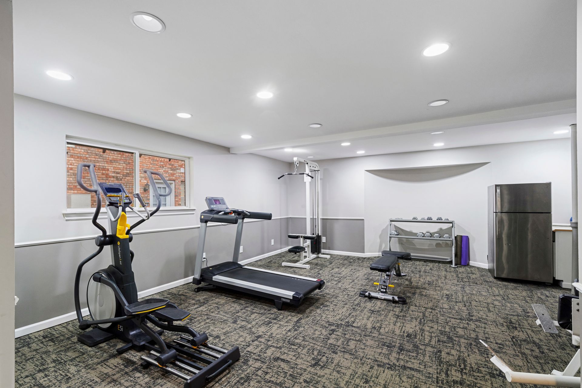 Gym with elliptical, treadmill, weights, bench, and refrigerator. Gray carpet, white walls, and bright ceiling lights.