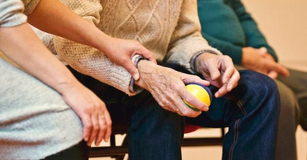 A woman is holding the hand of an elderly man while sitting in a chair.