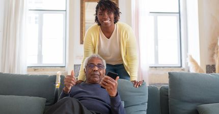 A woman is standing next to an older man sitting on a couch.