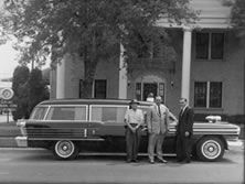 Black hearse with three men standing in front of a white columned building.