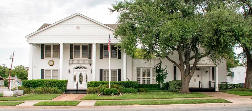 White two-story building with columns and an American flag; set on a green lawn with trees.