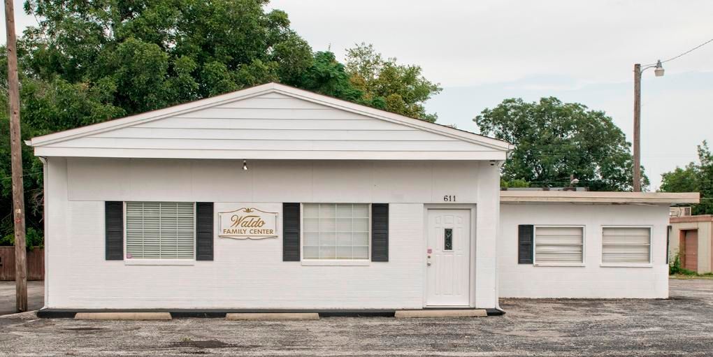 White building with black shutters and a small sign, set against a backdrop of trees and a cloudy sky.