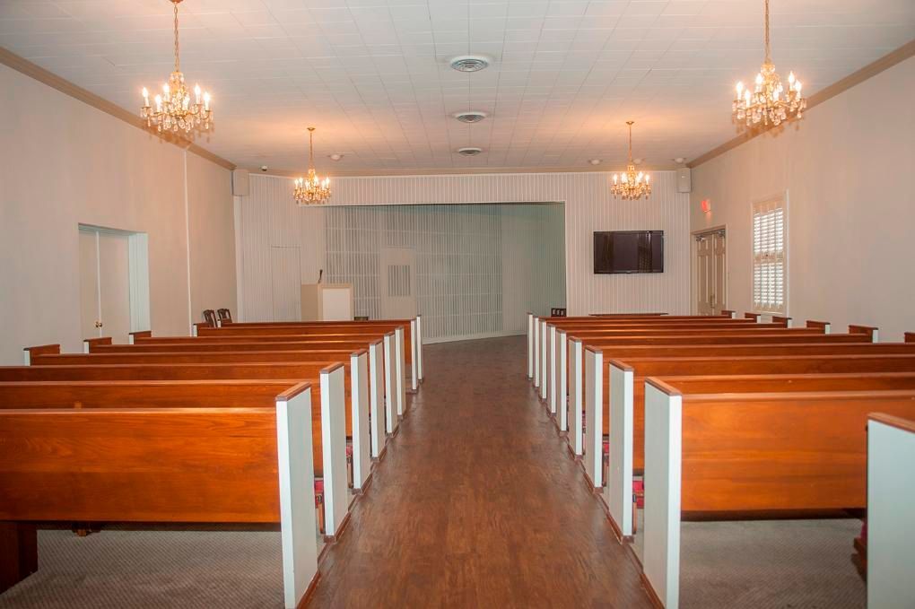 Empty church interior with rows of wooden pews, chandeliers, and a stage area.