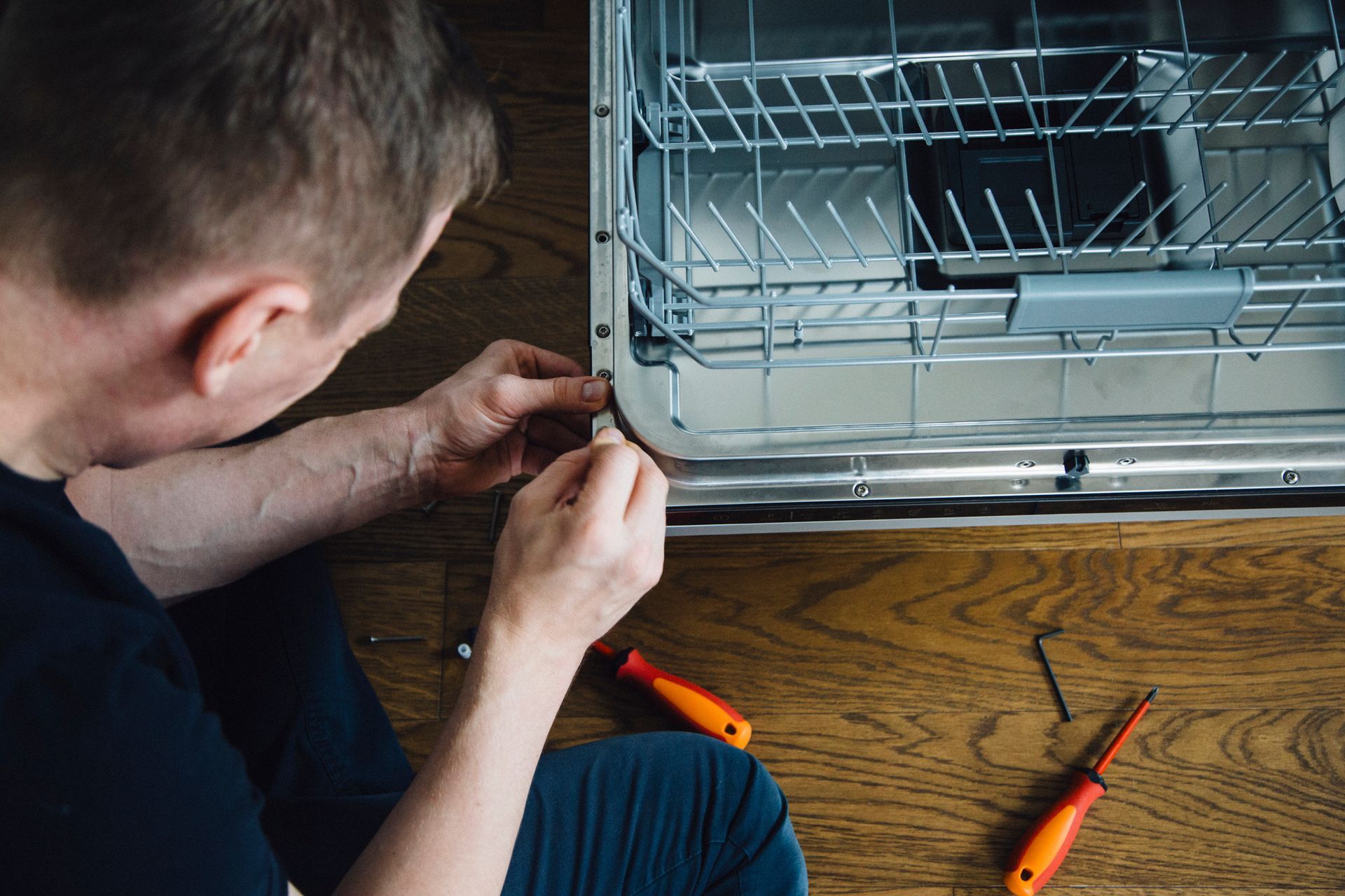 Person repairing a dishwasher on a wooden floor, using tools like screwdrivers.