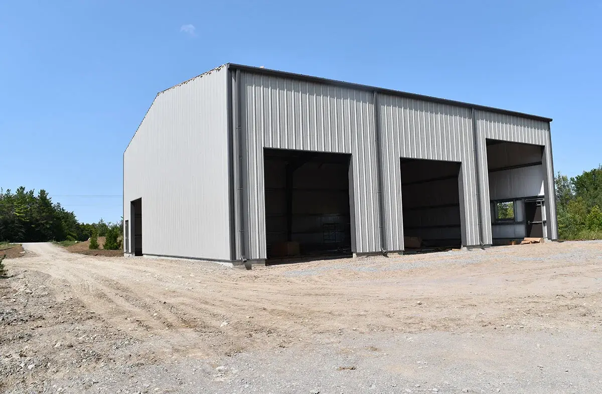 A large metal building with three garage doors is sitting on top of a dirt road.
