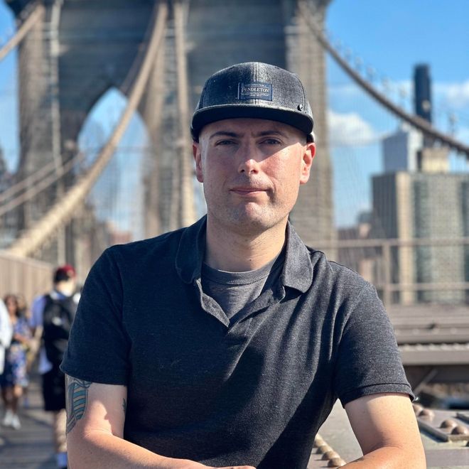Man wearing a cap and dark shirt, posing on the Brooklyn Bridge, looking at the camera.