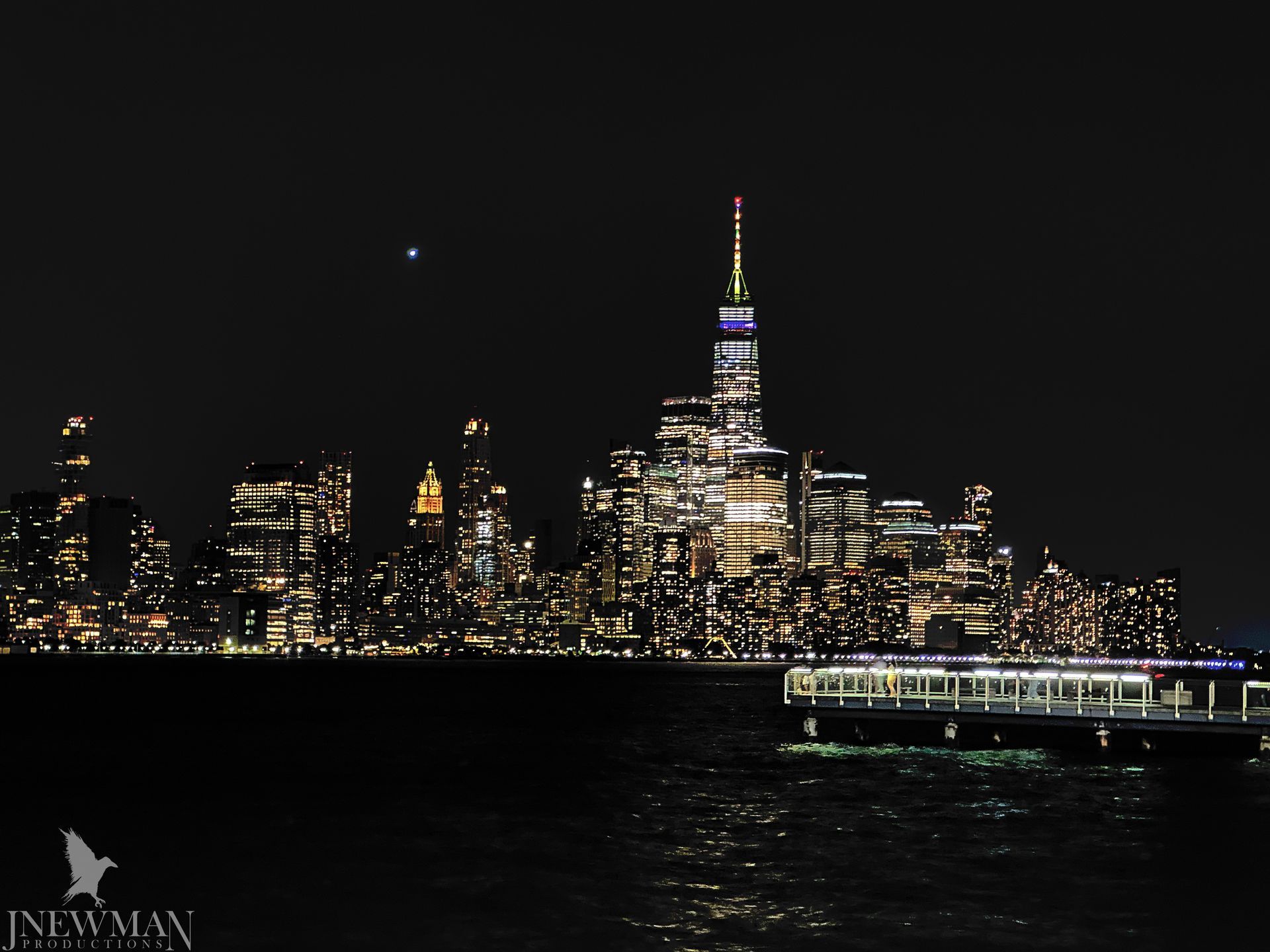 New York City skyline at night with illuminated buildings and water in the foreground.