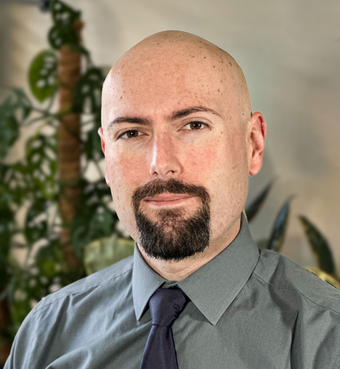 Man with bald head and goatee wearing a grey shirt and dark blue tie.