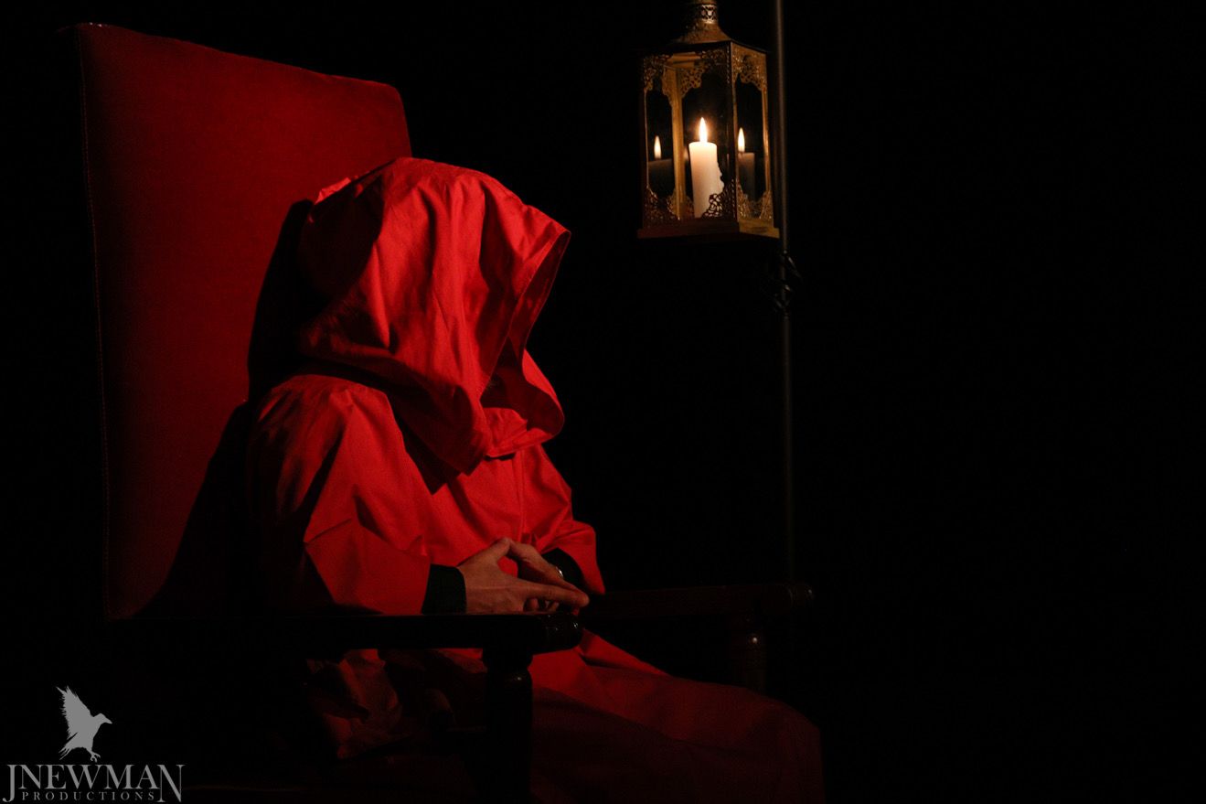 Person in red hooded robe seated in dimly lit room, holding hands, candle lit lantern.
