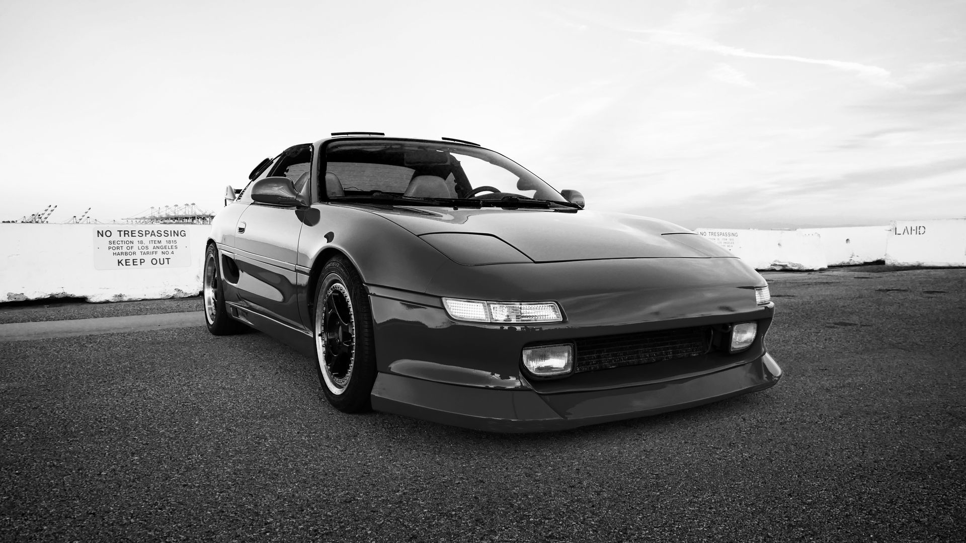 Black and white image of a sporty black Toyota MR2 car parked on a paved surface, front view.