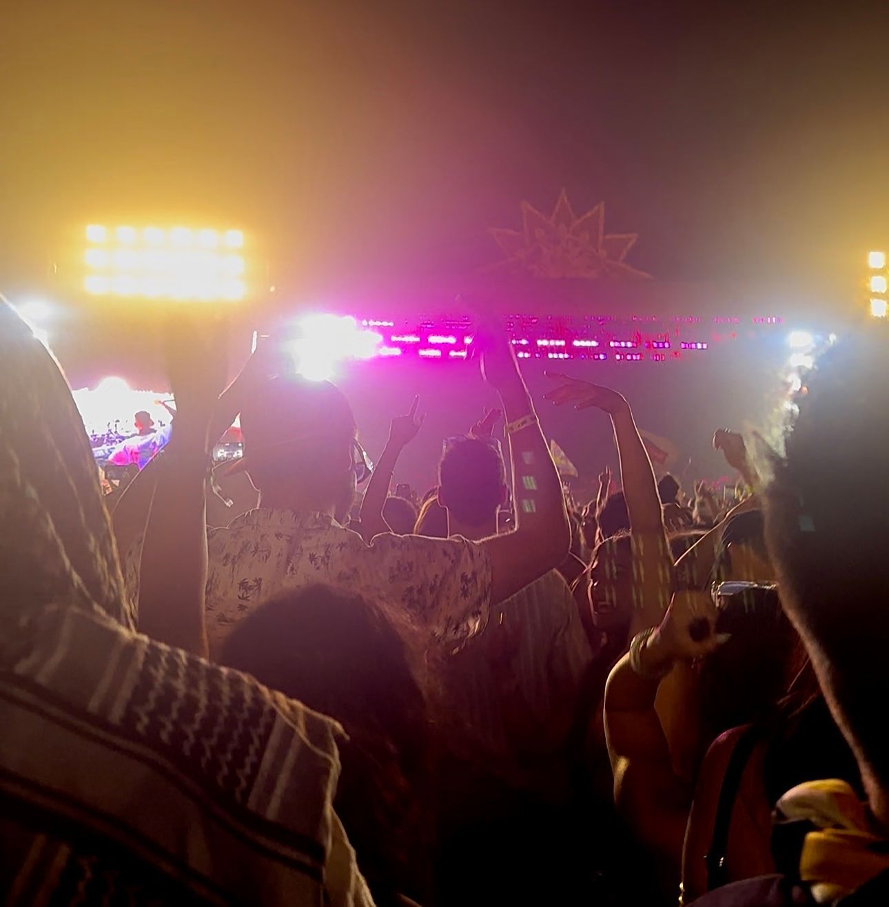 Concert crowd with arms raised toward a stage illuminated by bright pink and yellow lights.
