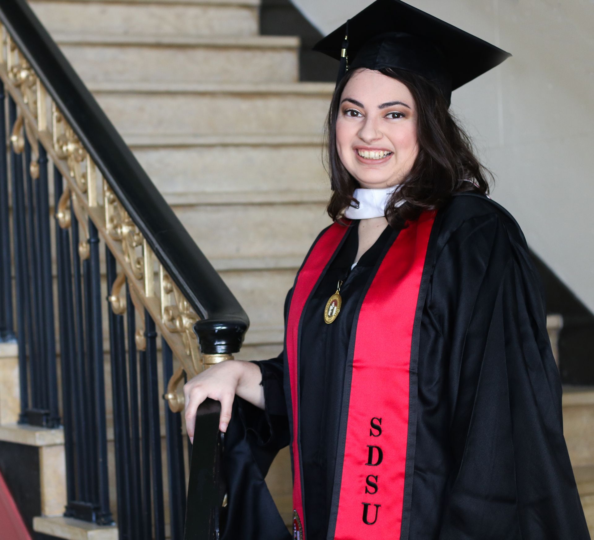 Woman in graduation attire smiles on a staircase, wearing a red and black sash with 