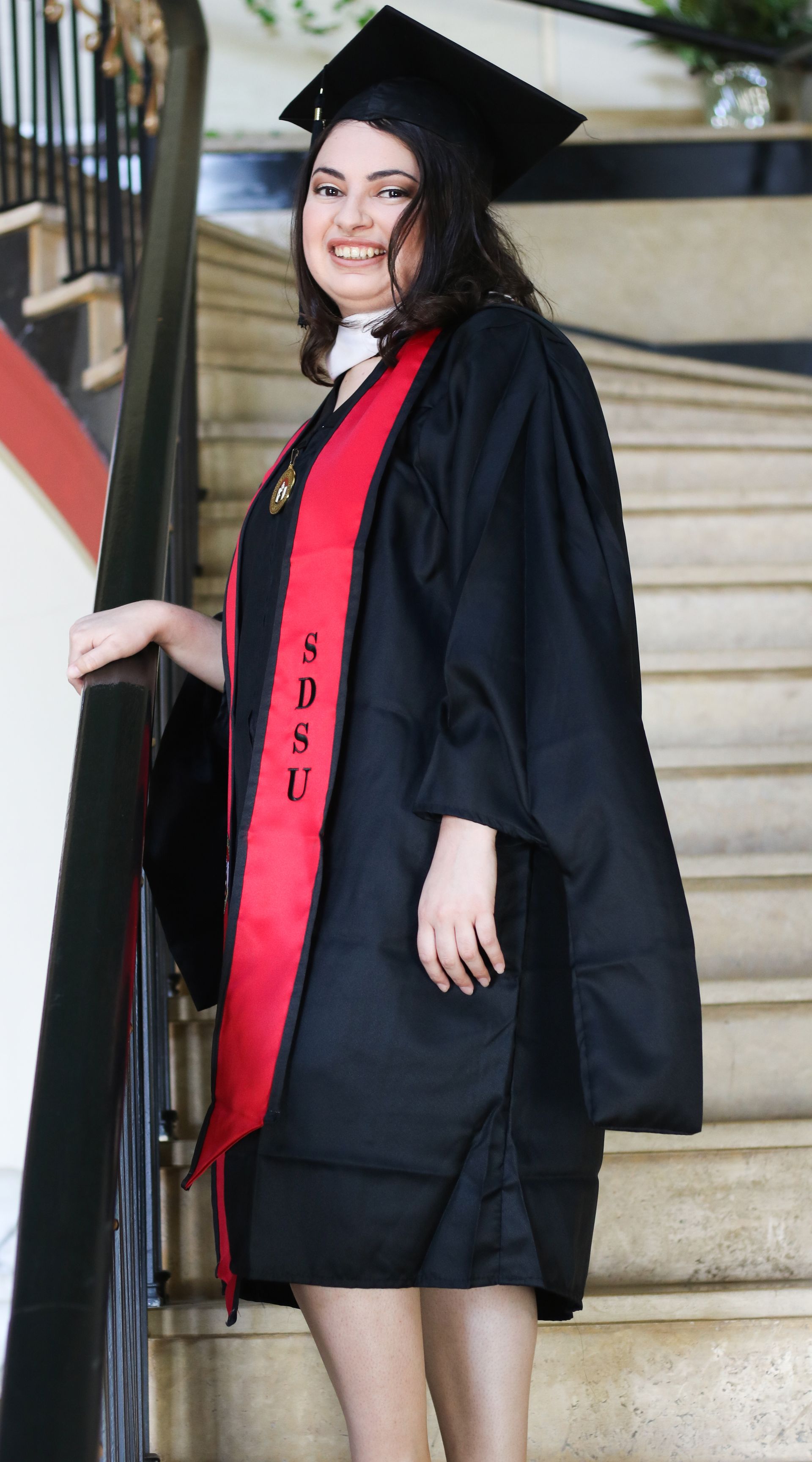 Woman in graduation cap and gown stands on stairs, smiling. Red sash displays 