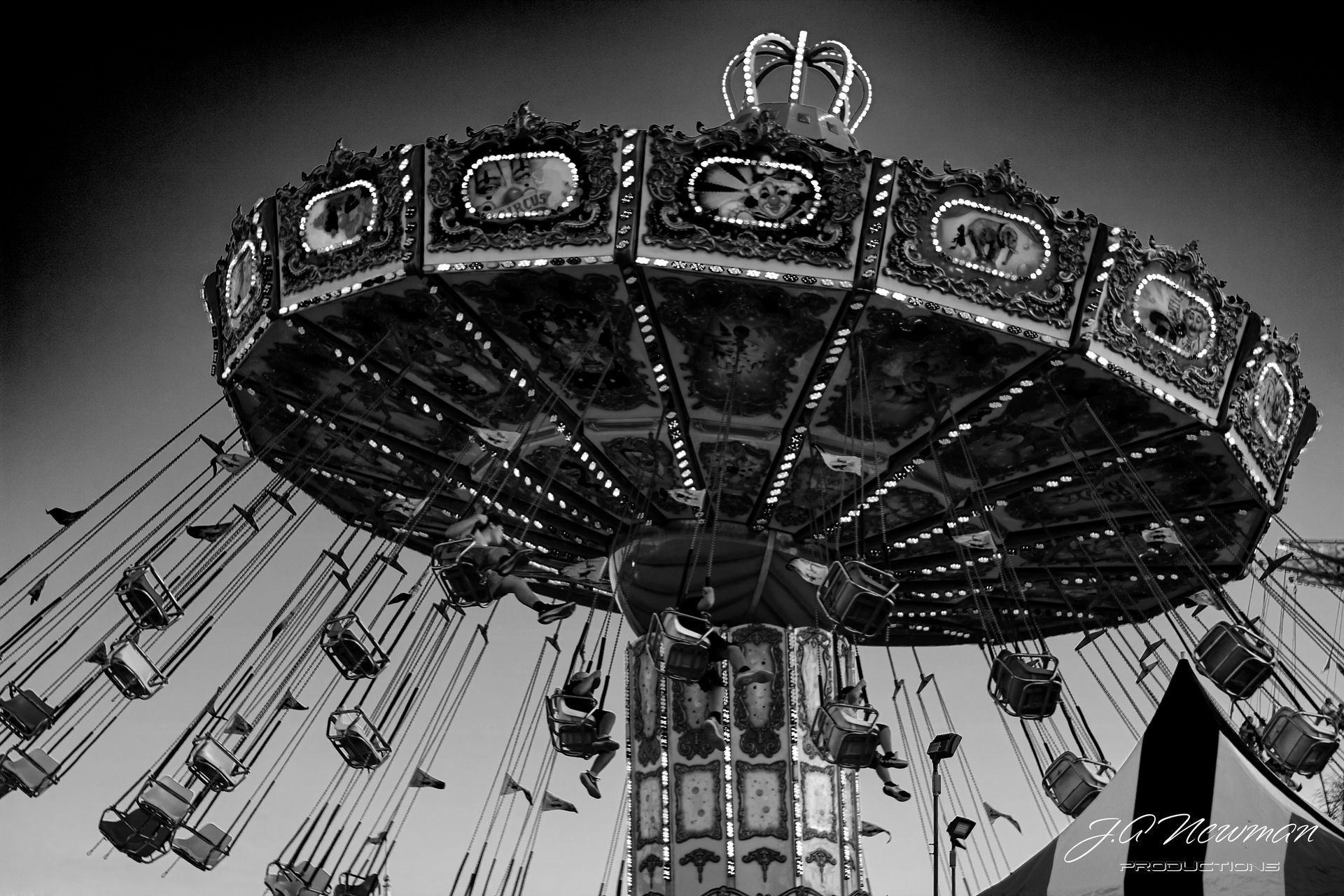 Black and white photo of a carnival swing ride, illuminated with lights, in motion with riders.