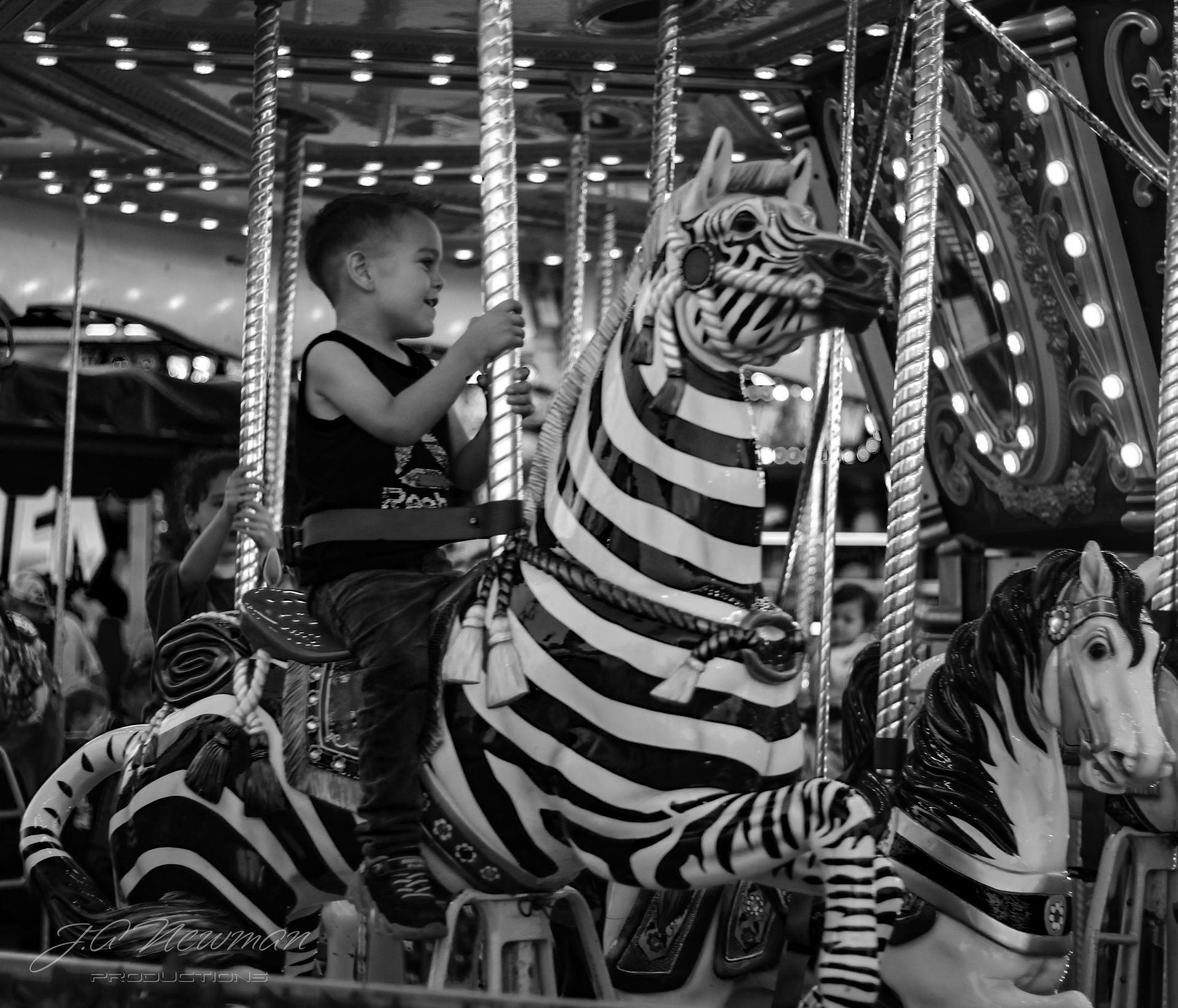 Boy on a zebra-striped carousel horse, smiling. Bright lights, other carousel figures and people in background.