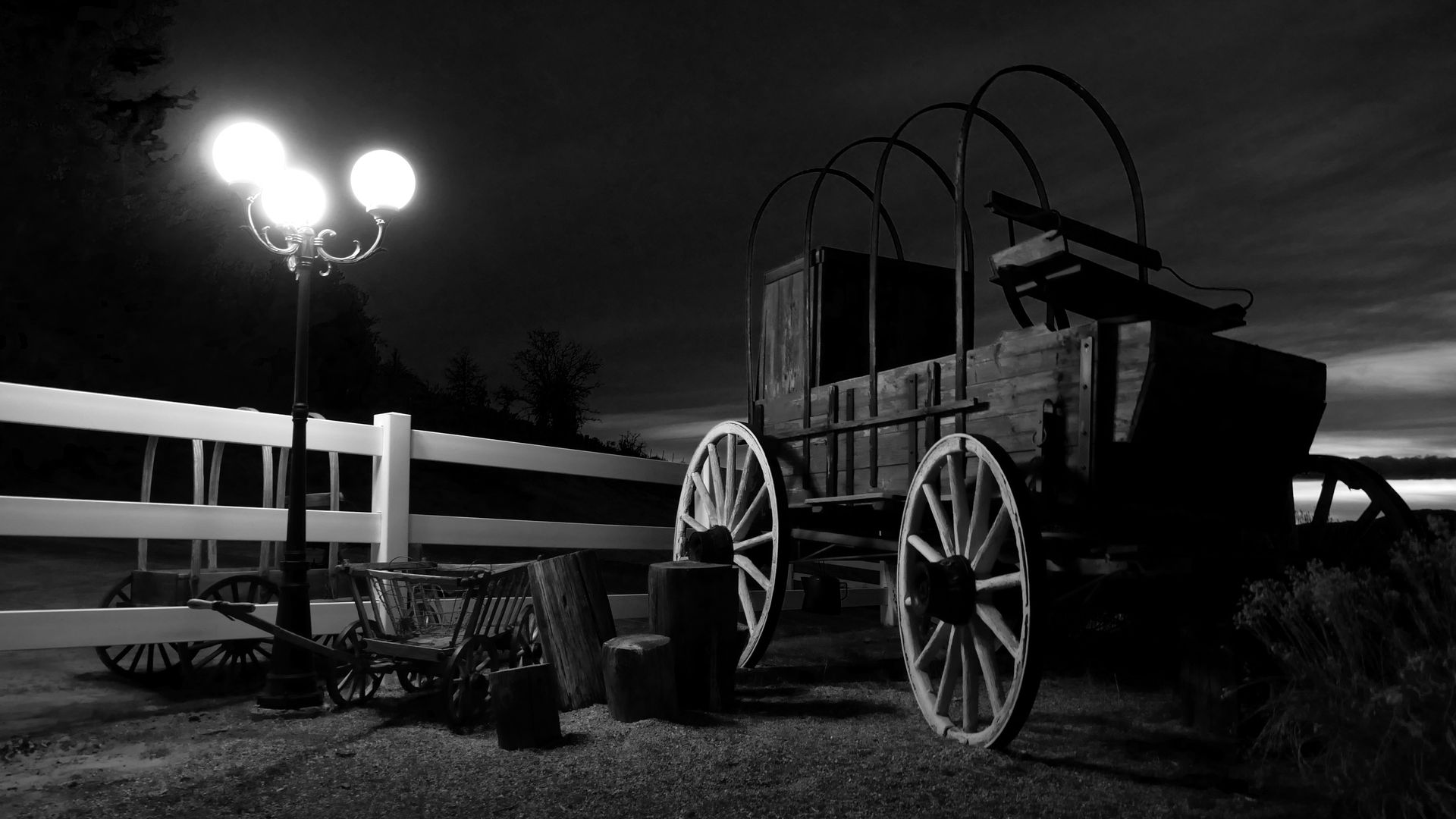 Black and white photo: a covered wagon next to a lit streetlamp and a white fence at night.