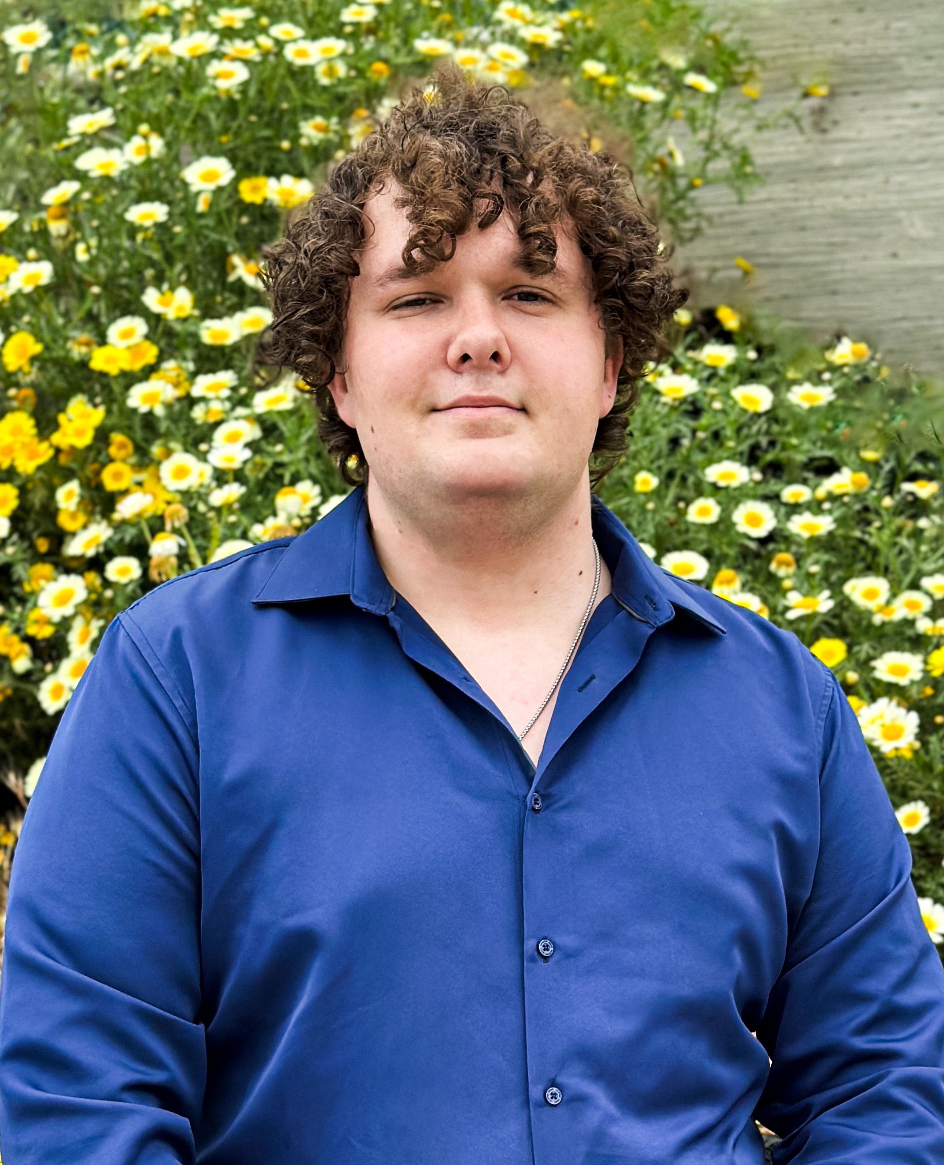 Man with curly brown hair wearing a blue button-up shirt, standing in front of yellow flowers.