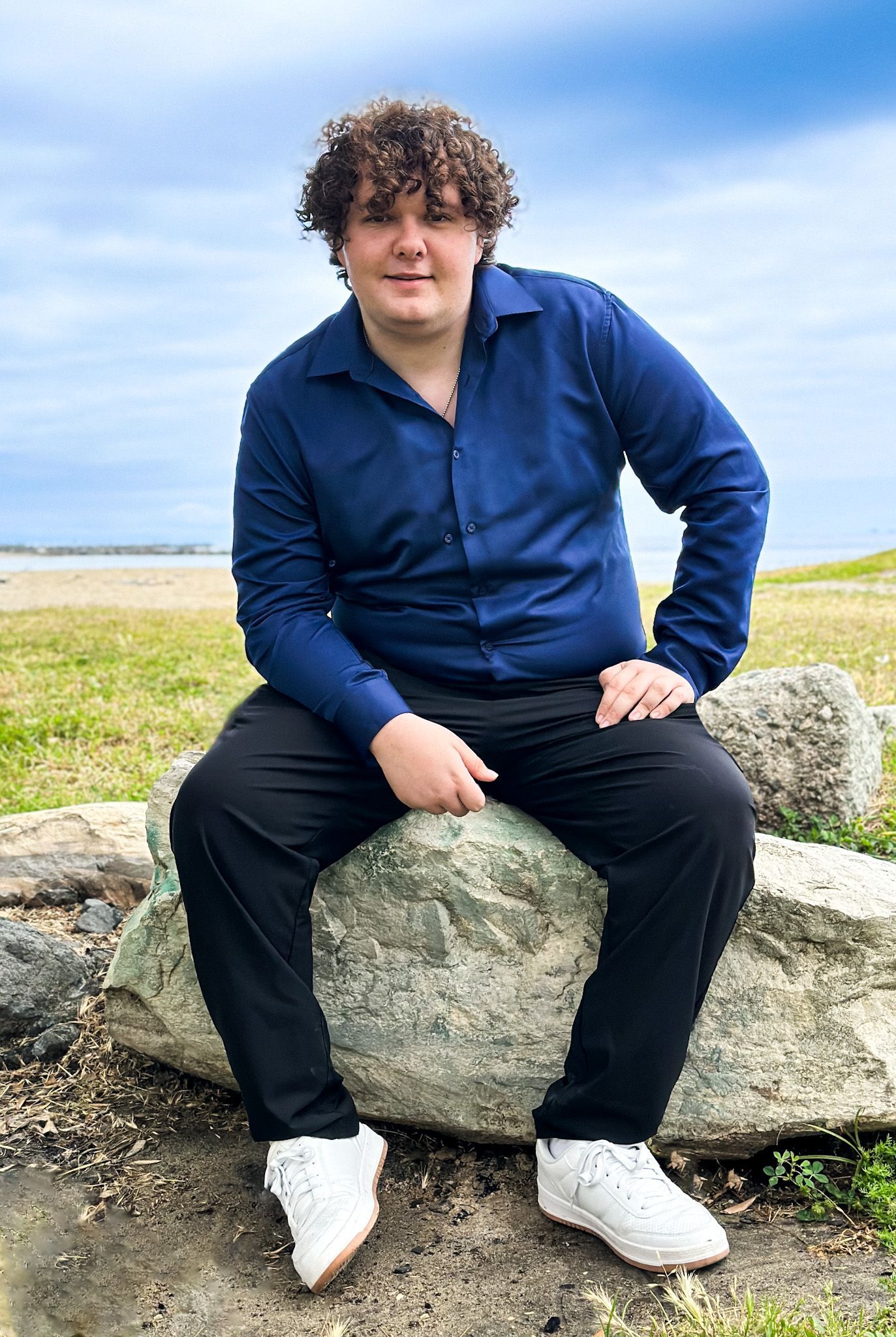 Person in navy blue shirt and black pants sits on a rock near a grassy area with a beach in the background.