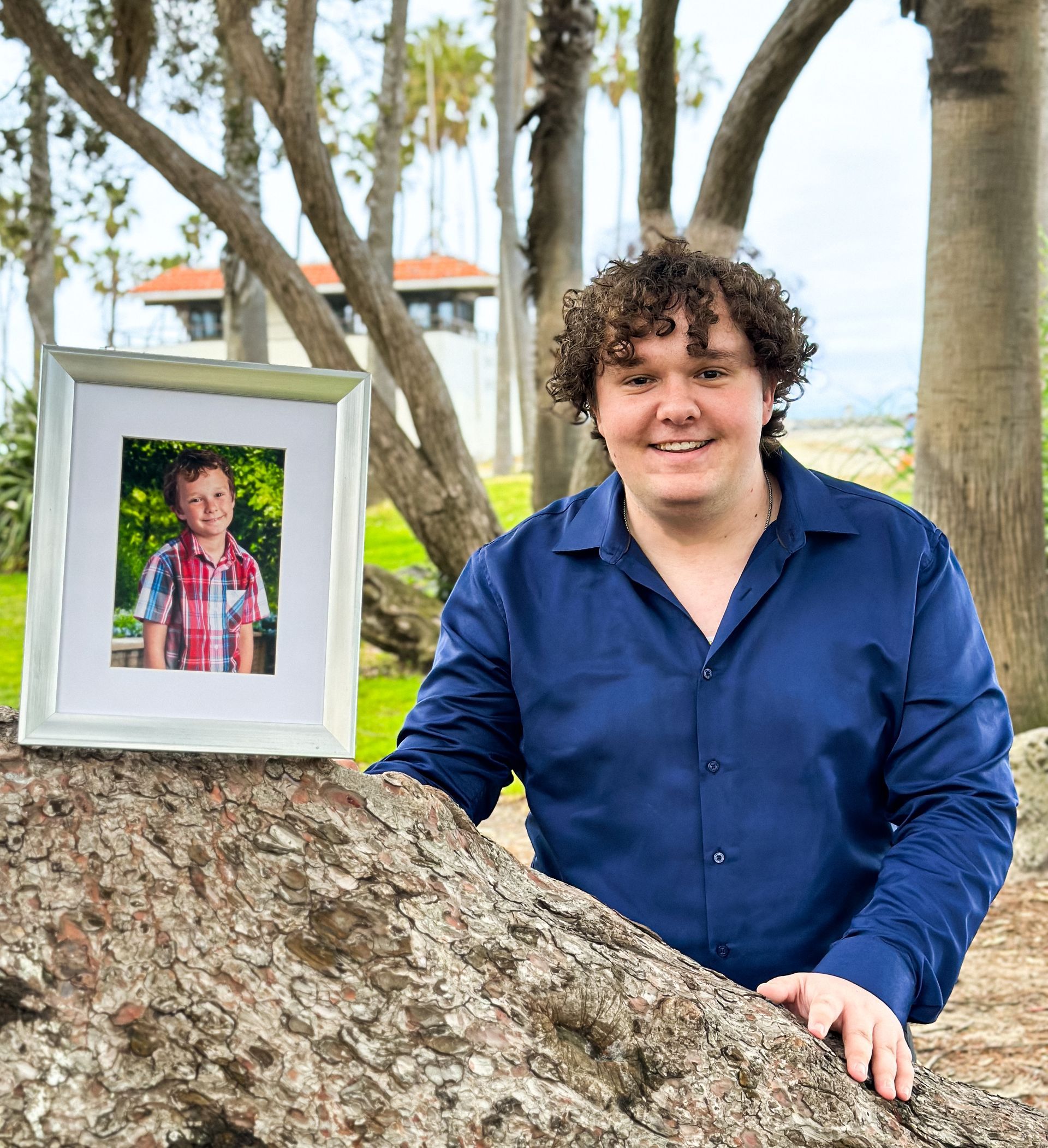 Man with curly hair smiles, beside a framed childhood photo in an outdoor setting.