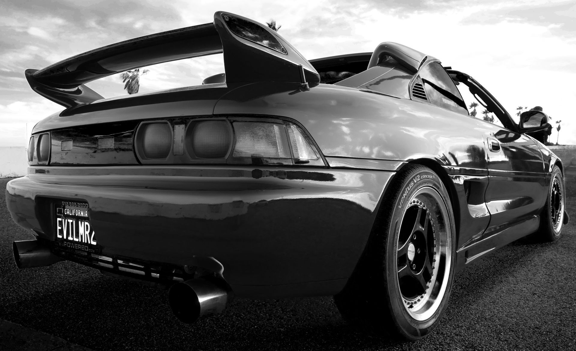 Black and white photo of a sports car's rear, with a spoiler, exhaust pipes, and black wheels on asphalt.