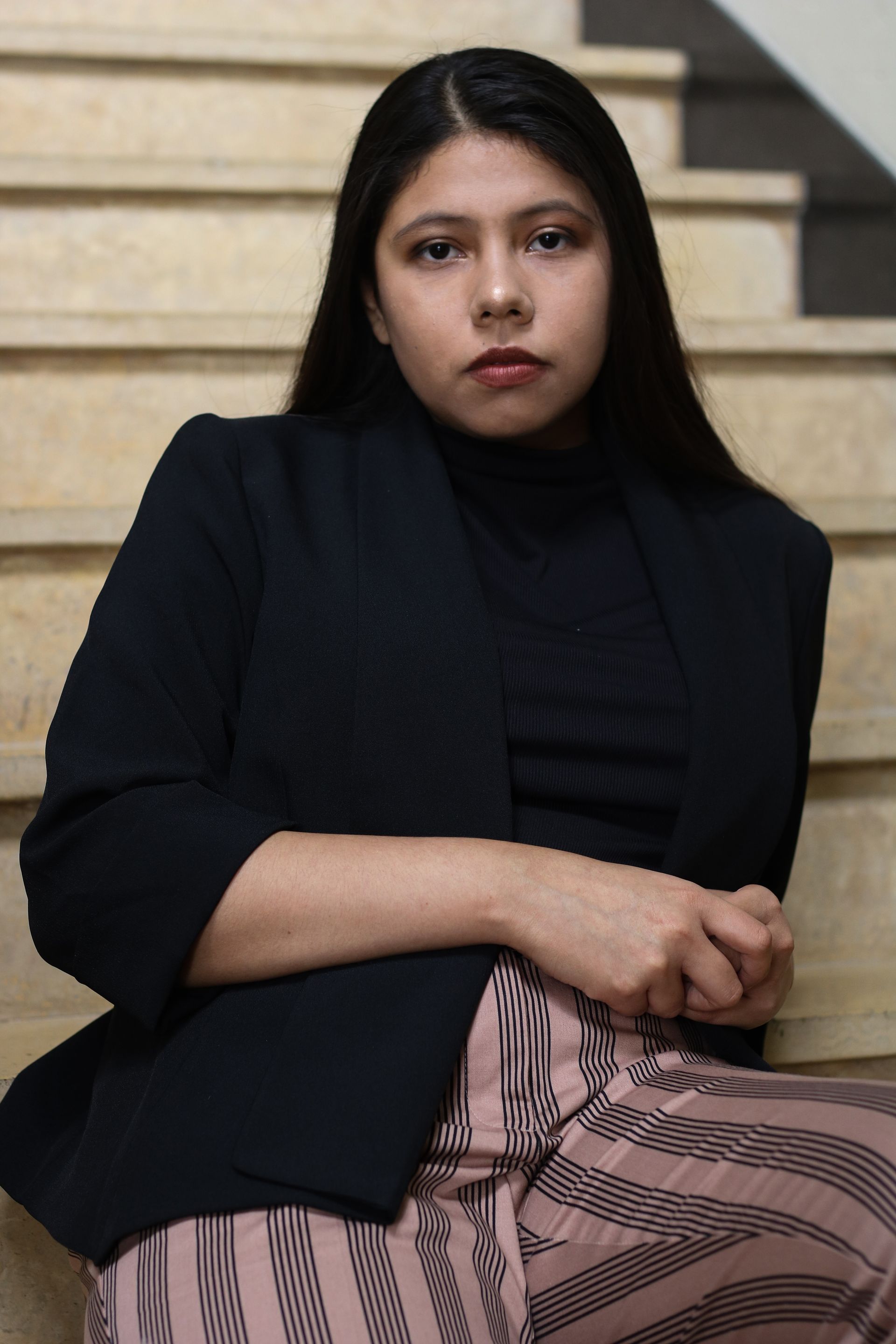 Woman in black blazer and pink striped pants sits on stairs, looking directly at the camera.