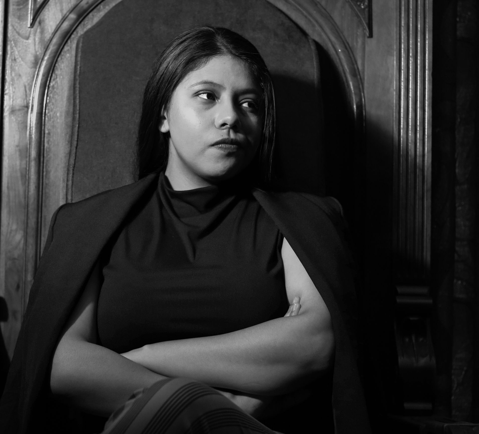 Woman in dark turtleneck and blazer, arms crossed, sits in ornately carved chair.