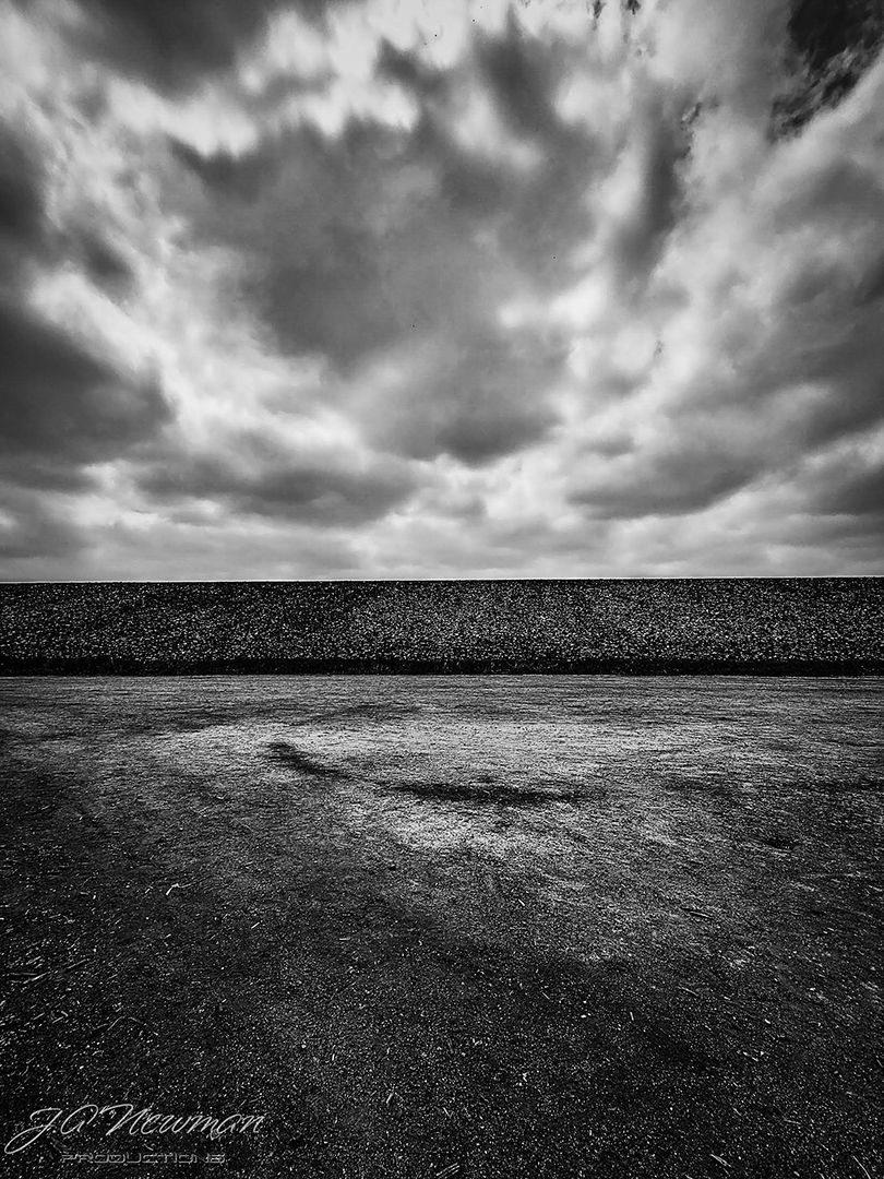 Black and white photo: Concrete ground leading to a textured wall under a cloudy sky.