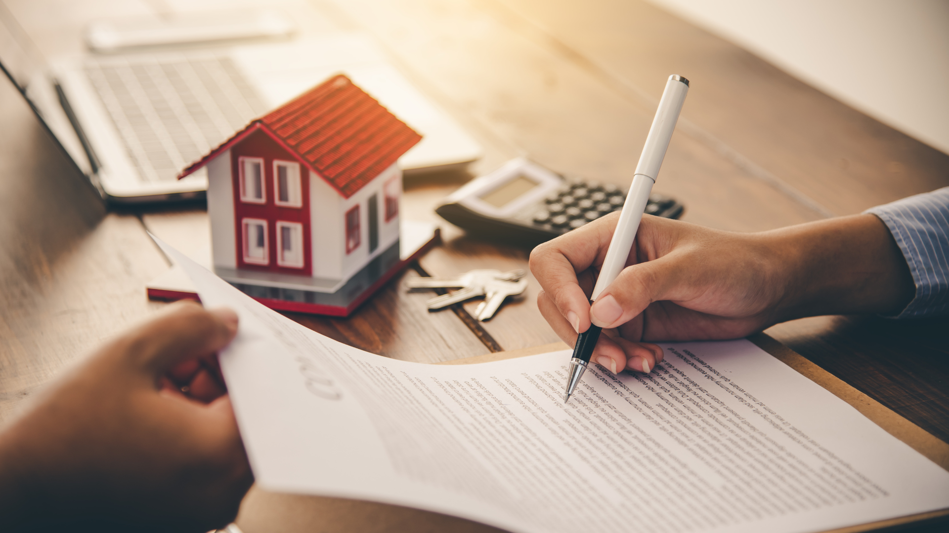 Person signing real estate contract; model house, keys, calculator, laptop on table.