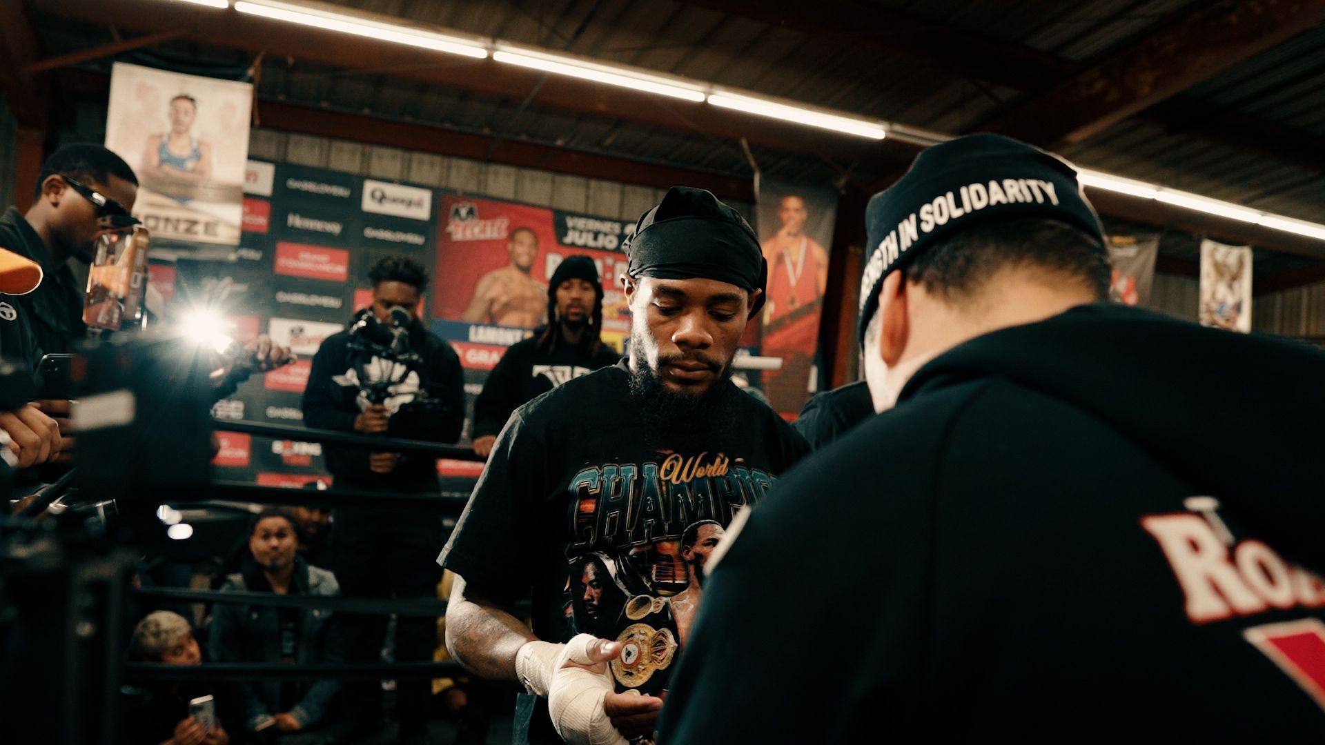 Lamont Roach Jr. wraps hands in a boxing ring, surrounded by people, in a gym with posters. Image captured by Mason Taylor. 