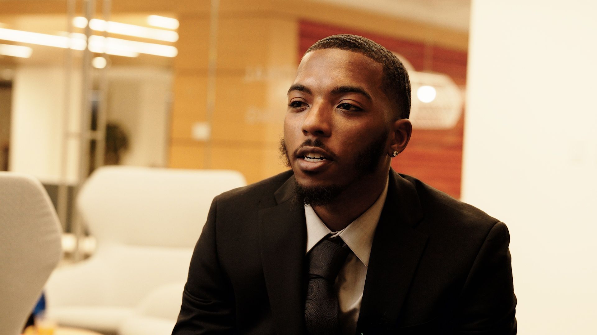 Man in black suit speaking, in an office setting.