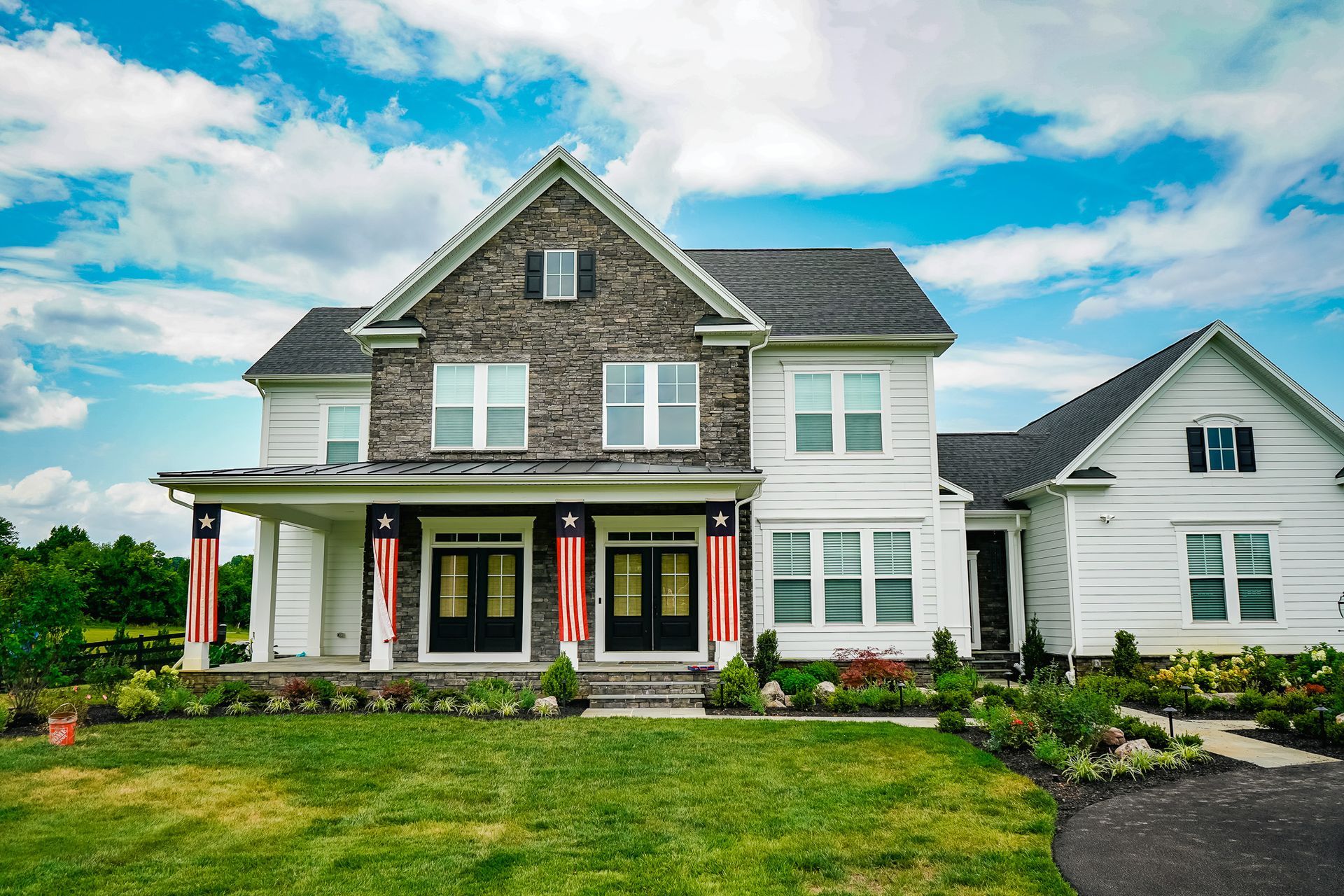 Two-story white house with stone accents, black doors, American flags, and green lawn under a blue sky.