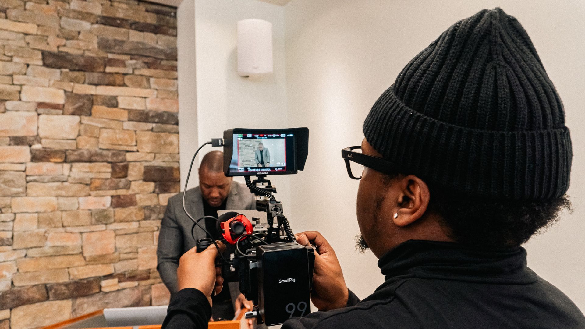 Person filming another person in a room with a stone wall; camera equipment is visible.