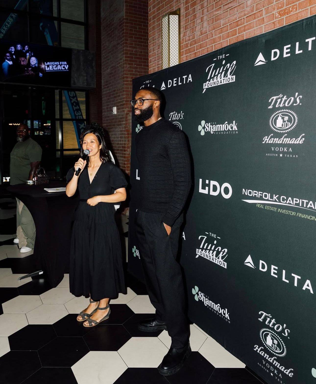 Woman speaking at a microphone next to a man. Event backdrop with company logos.