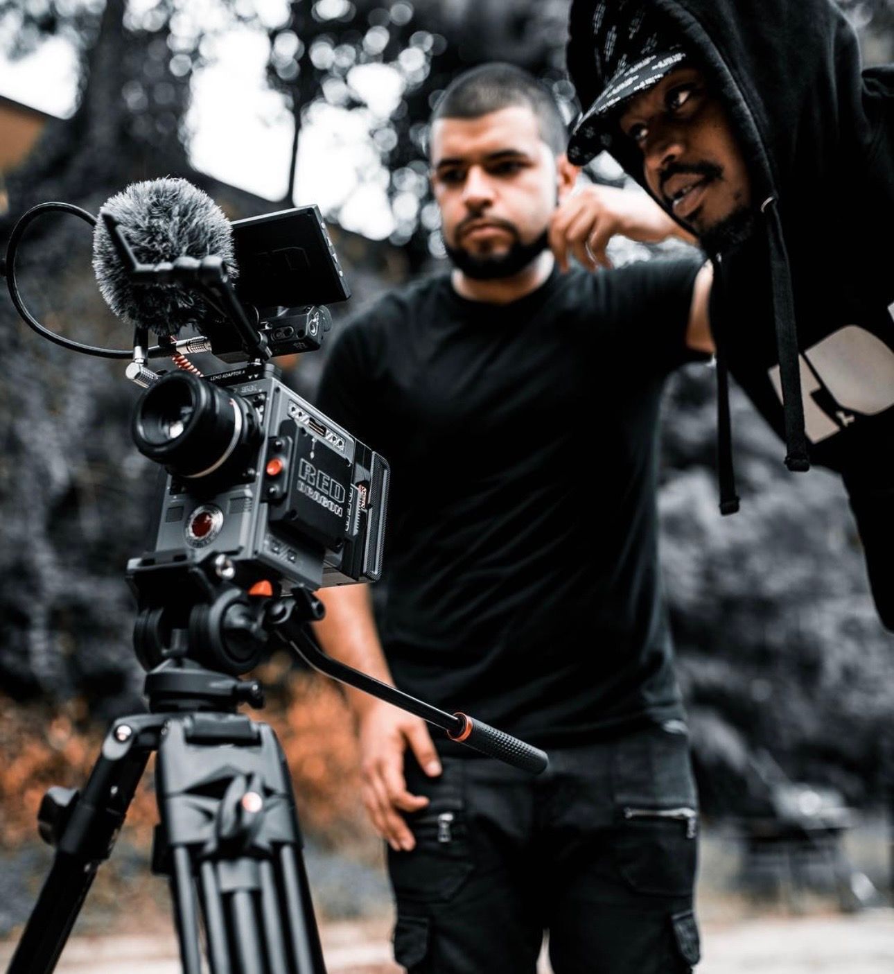Two people beside a video camera on a tripod, one pointing, the other looking. Outdoors, muted tones.
