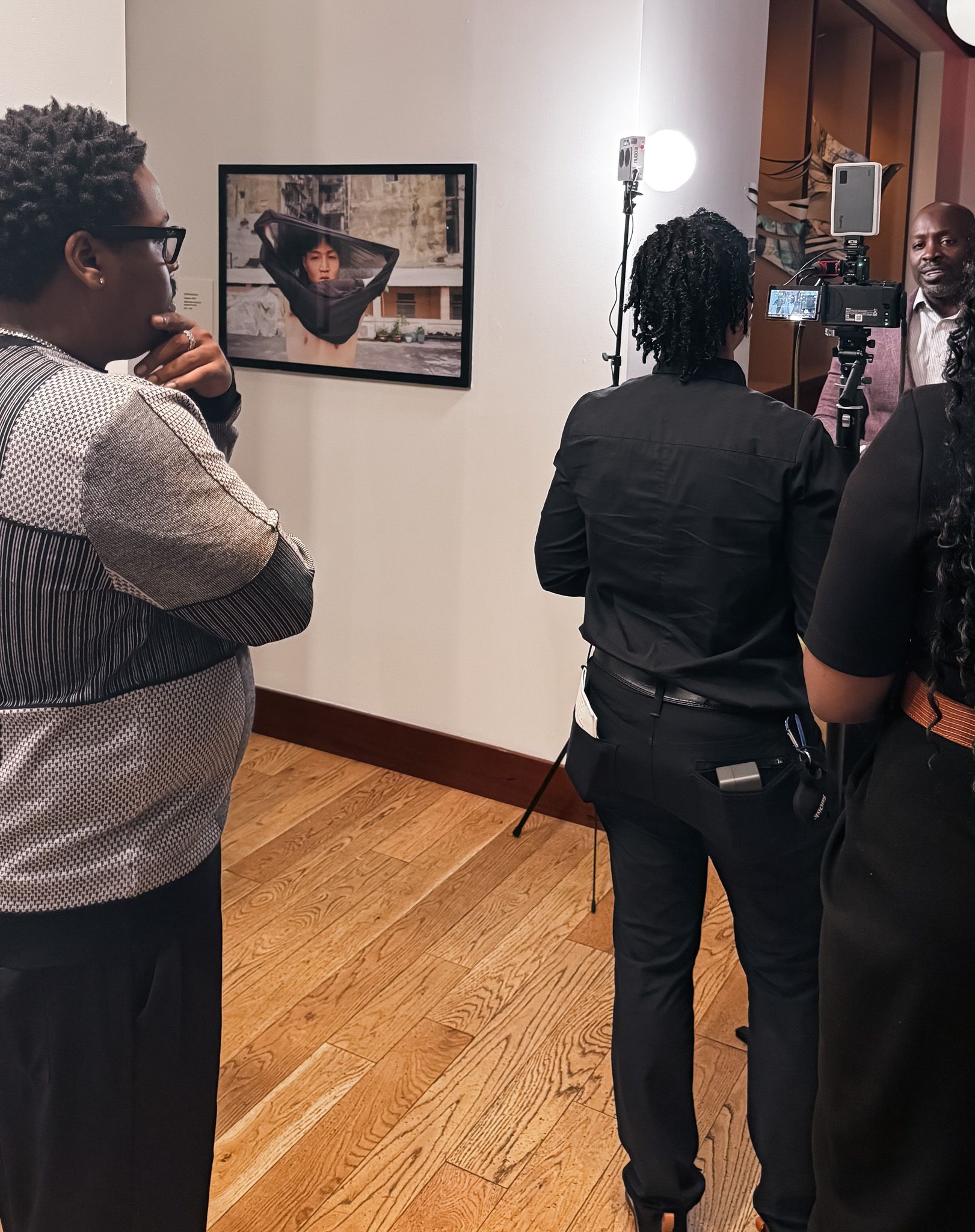 People gather; filming setup. Artwork hangs on the wall. Wooden floor, neutral walls, natural light.