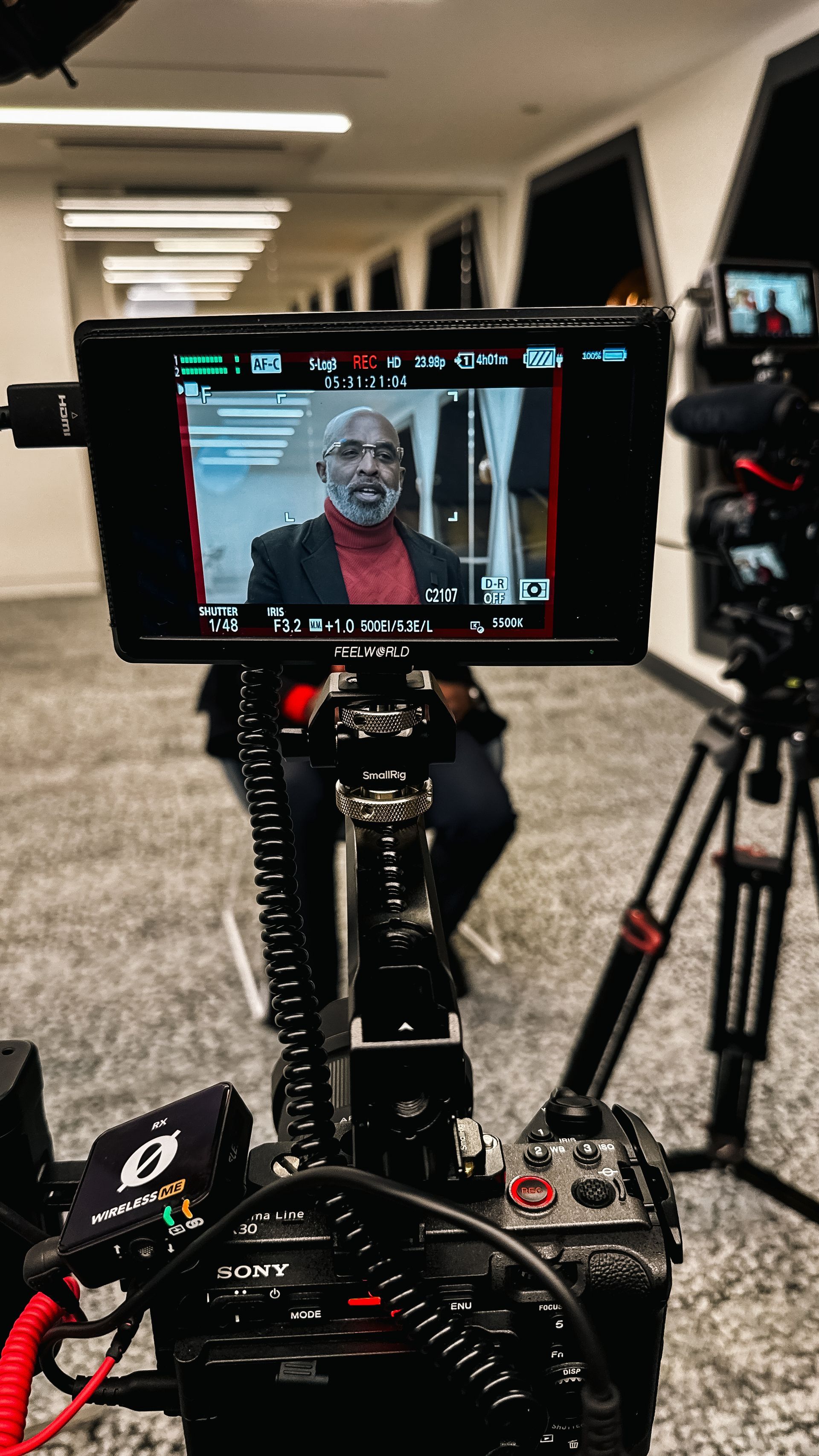 Video camera filming a man in a red shirt and blazer, indoors. Camera equipment in the foreground.