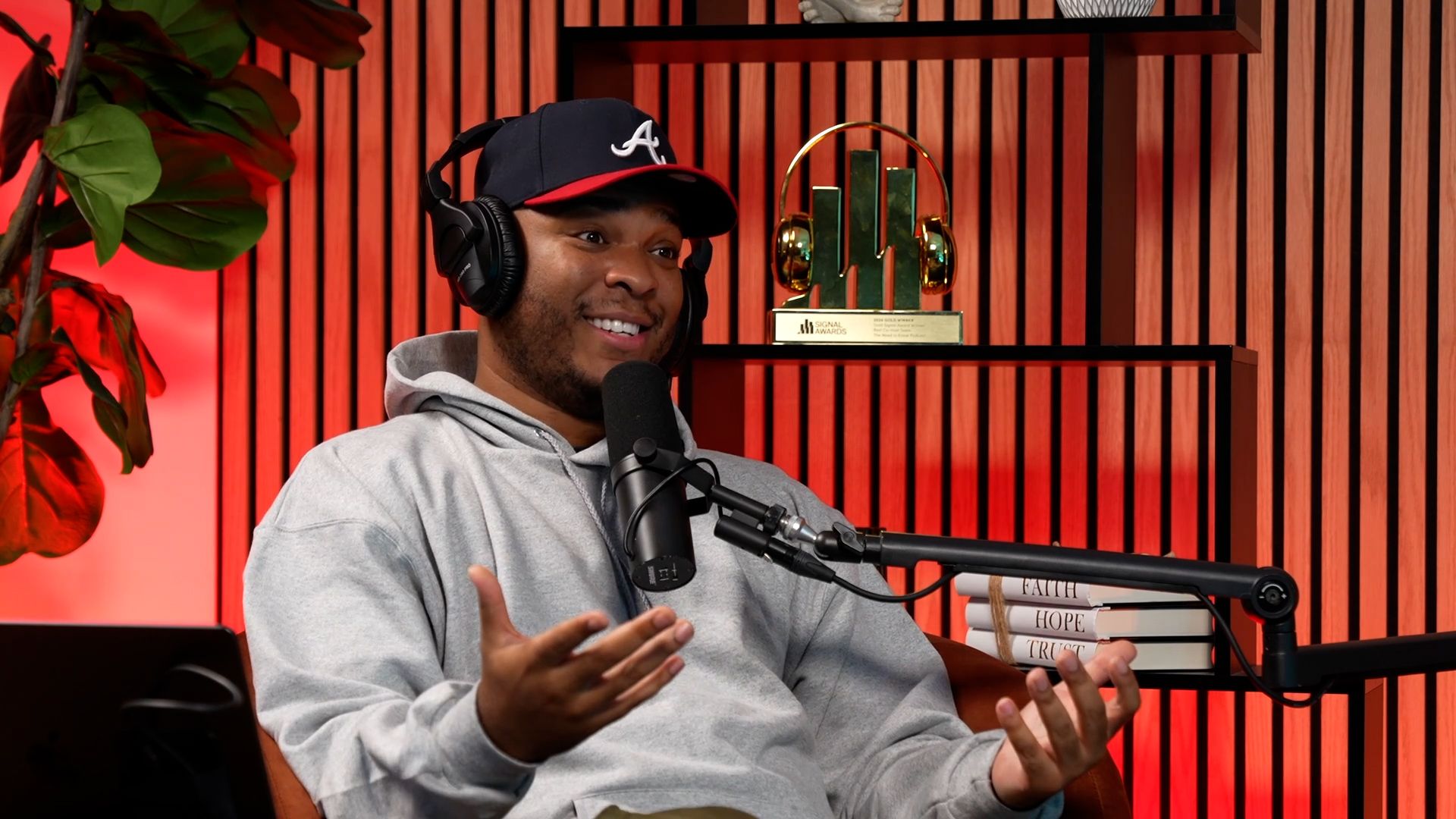 Man wearing headphones and a cap gestures while speaking into a microphone in a podcast studio.