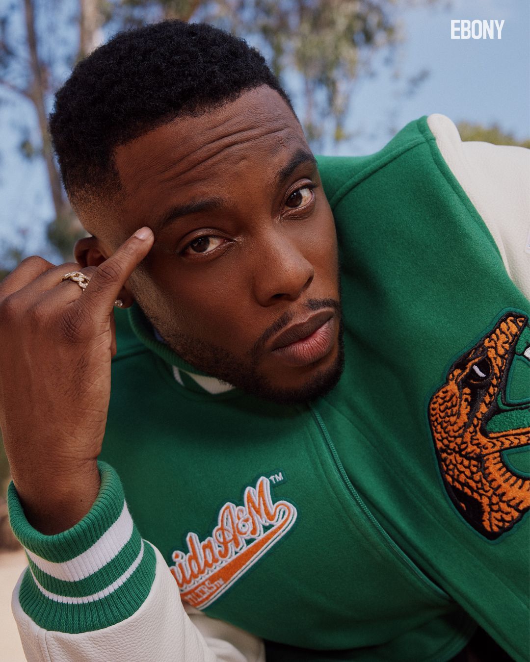 Woody McClain in a green and white jacket touches his temple, looking thoughtful. Outdoor setting.